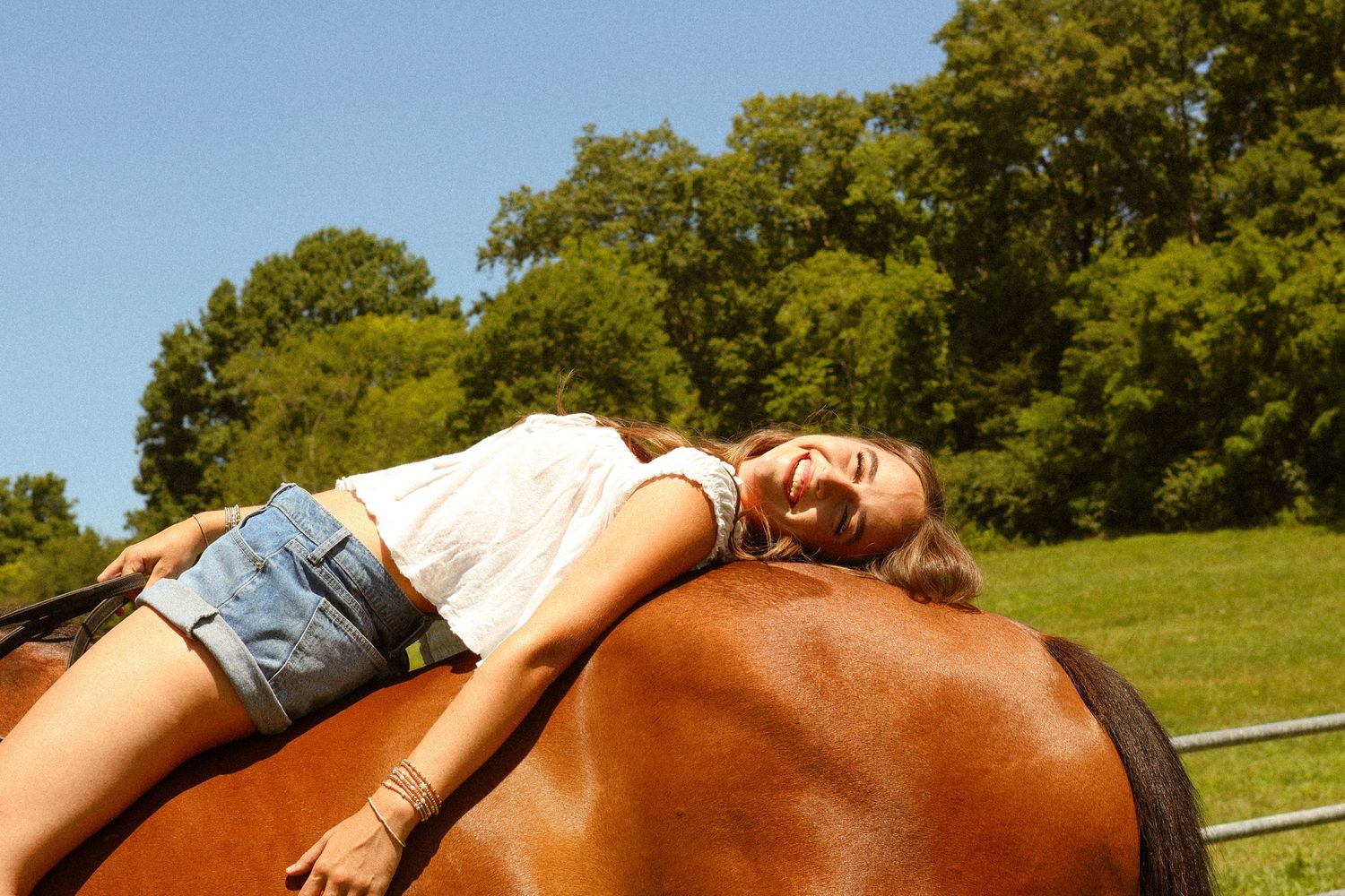 Young girl joyfully lying on a brown horse in a sunny green pasture, smiling with eyes closed.