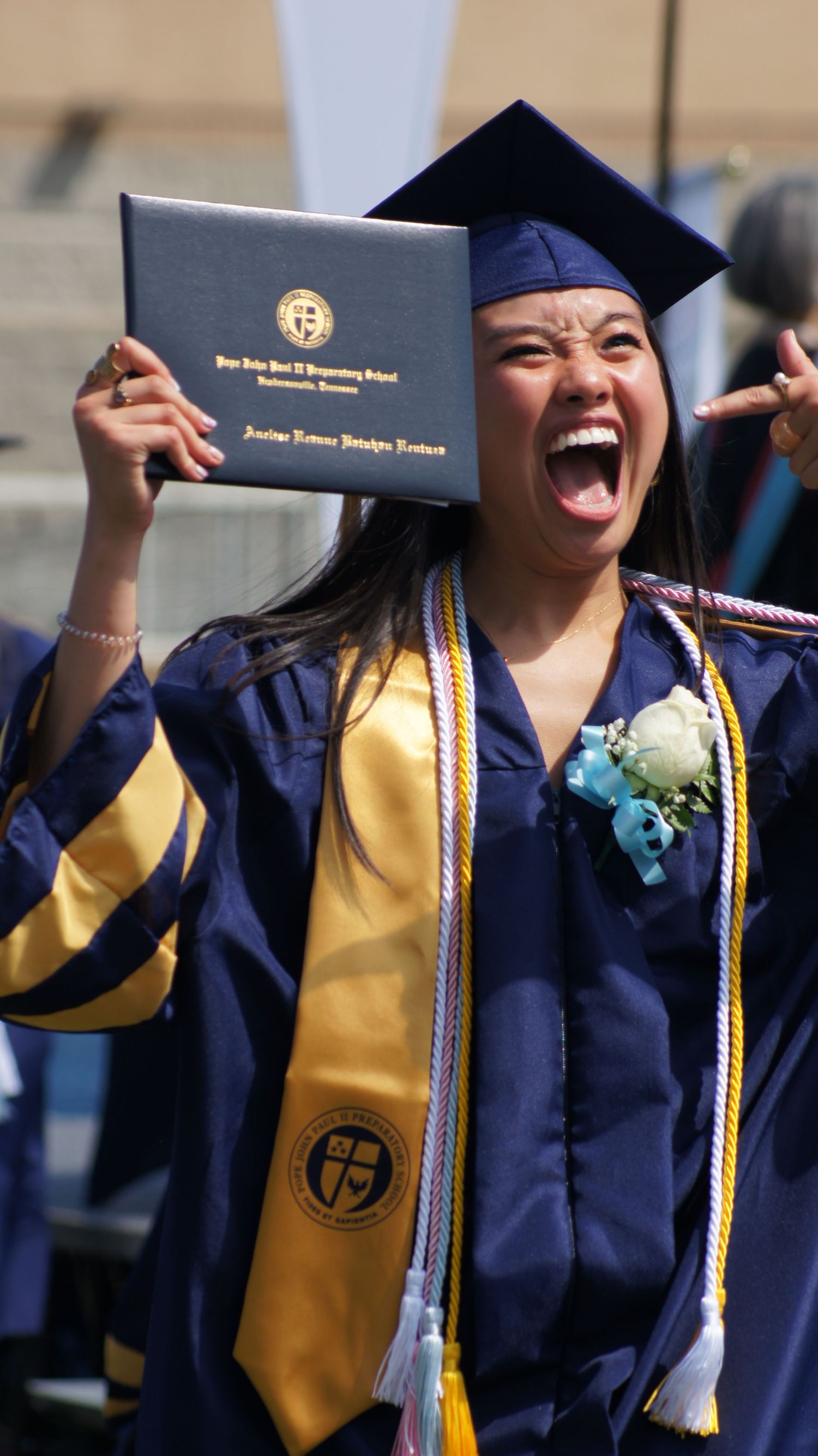 Graduate in blue and gold cap and gown celebrates joyfully while holding up diploma at commencement ceremony.