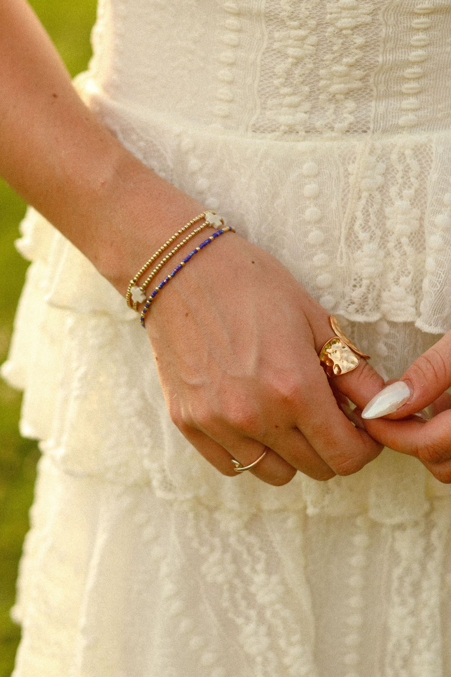 Close-up of a woman's hands wearing gold rings and a blue beaded bracelet, dressed in a white lace tiered dress.