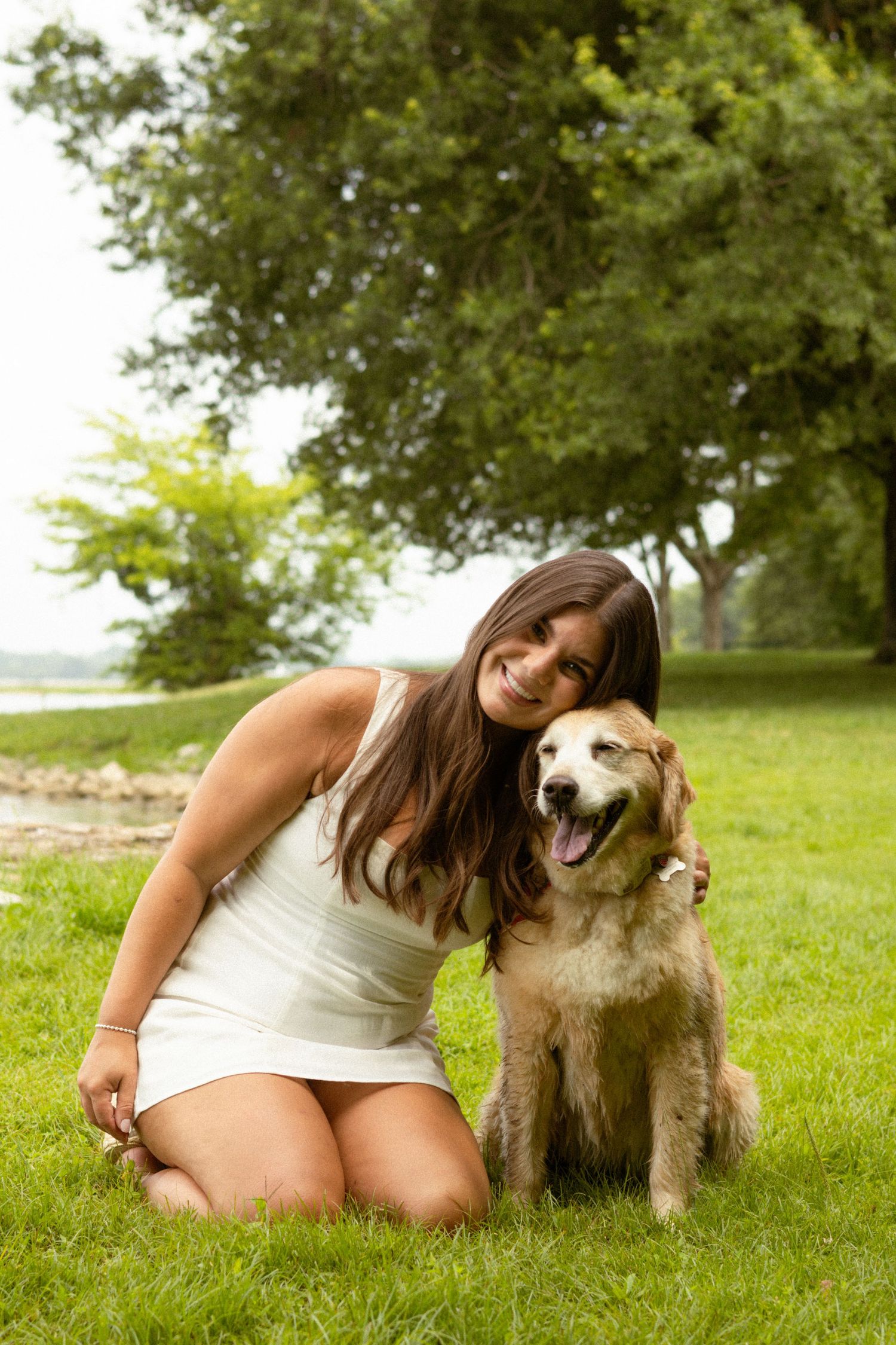 Young woman in white dress smiling and hugging her golden retriever dog on green grass in a park.