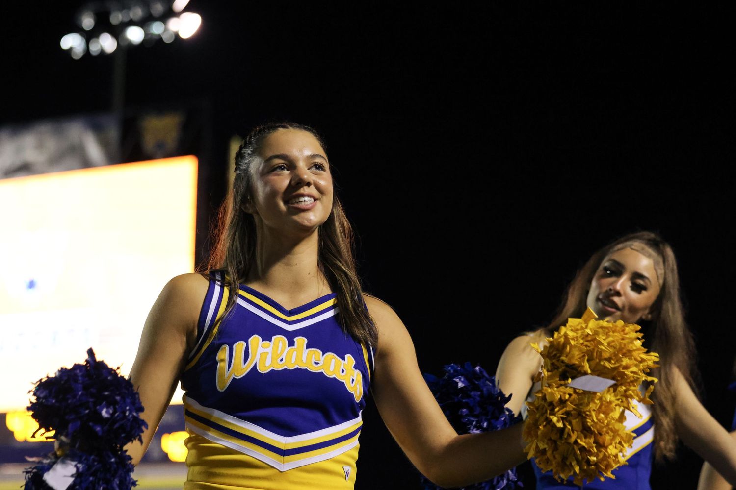 Cheerleaders in blue and yellow uniforms perform enthusiastically during a nighttime event.