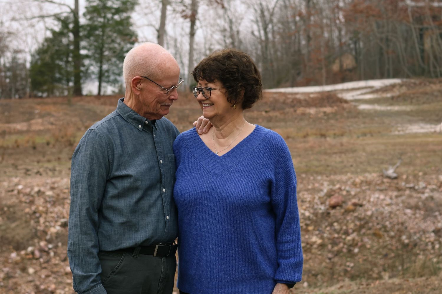 A couple in casual attire shares a tender moment outdoors during autumn with trees in the background.