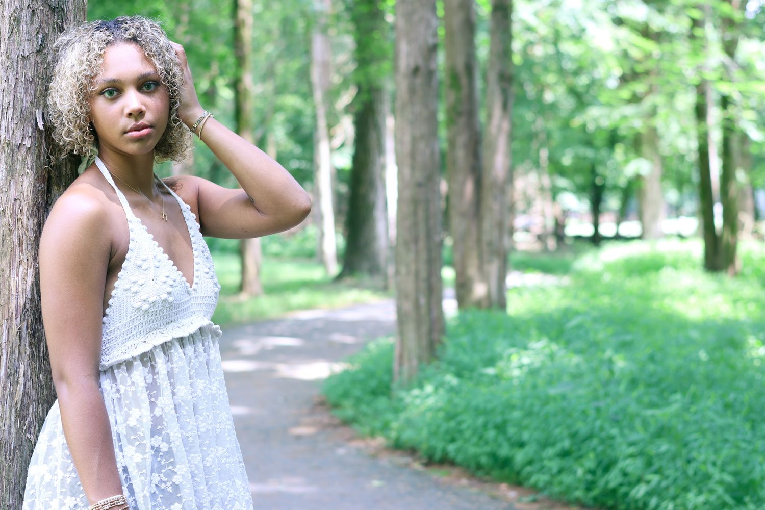 Person in white lace top poses along wooded path with natural lighting.