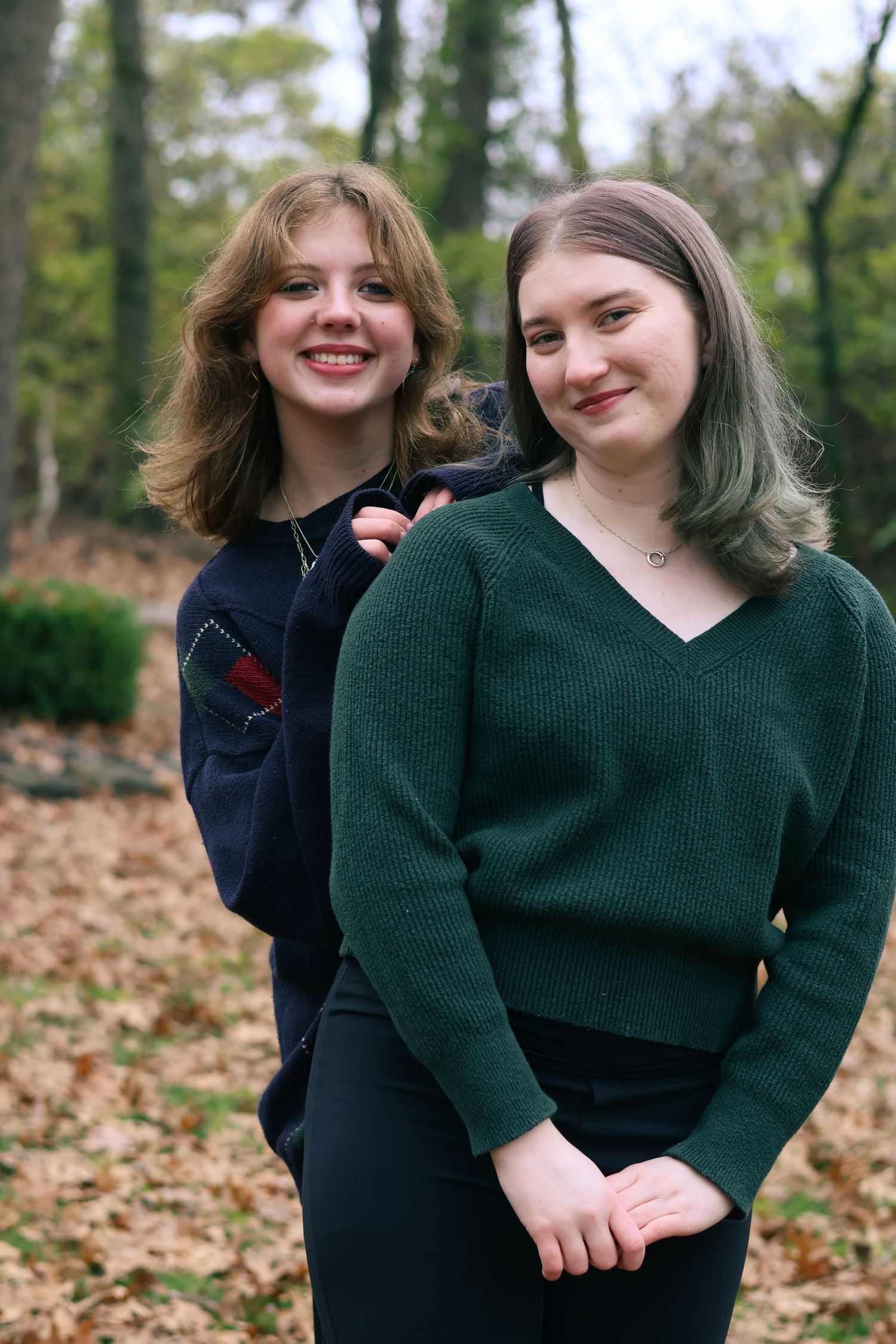 Two young women in sweaters smile together while standing in a wooded area with fallen autumn leaves.