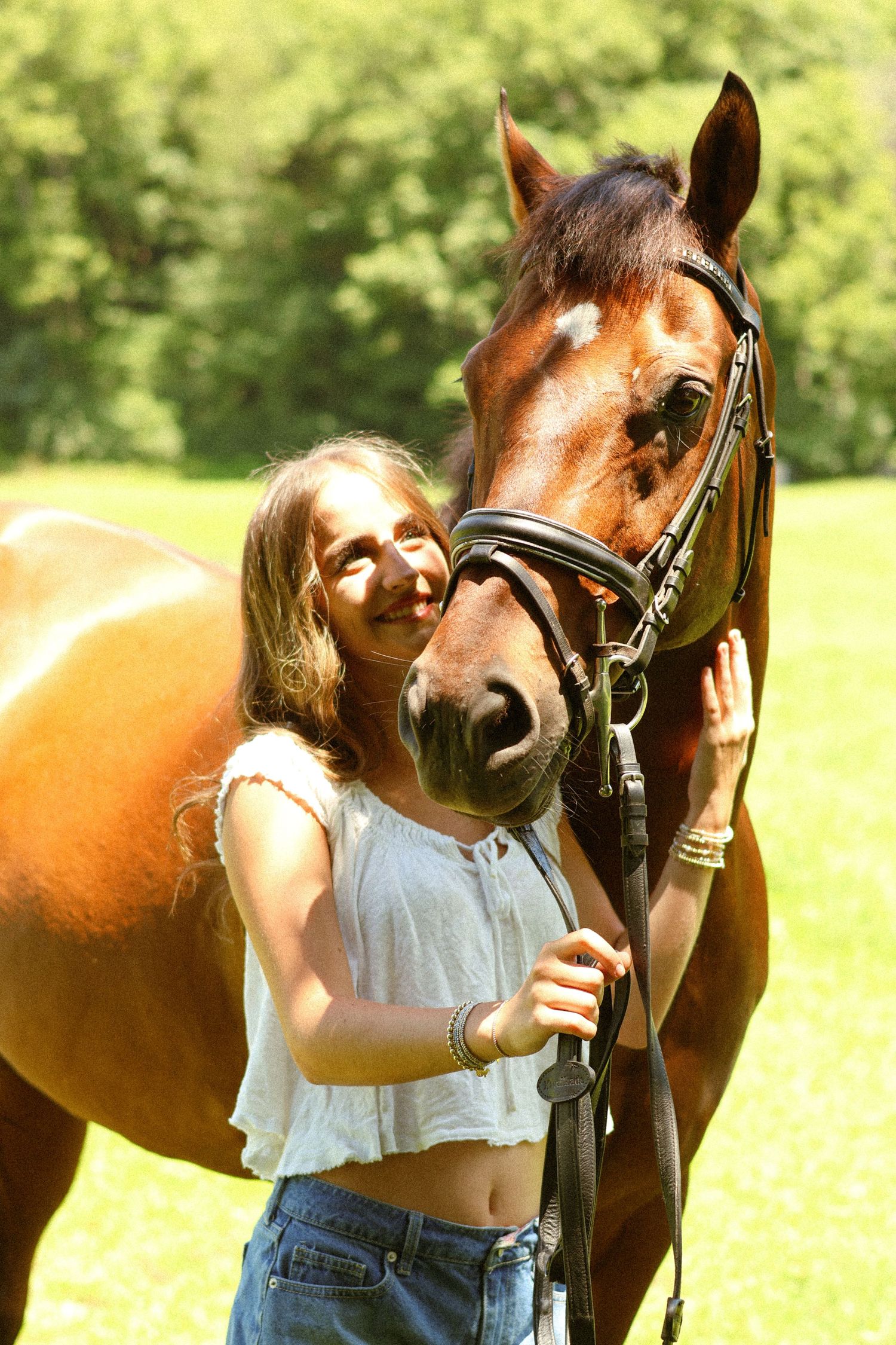 Young woman smiling and bonding with a brown bay horse outdoors in a sunny green field.