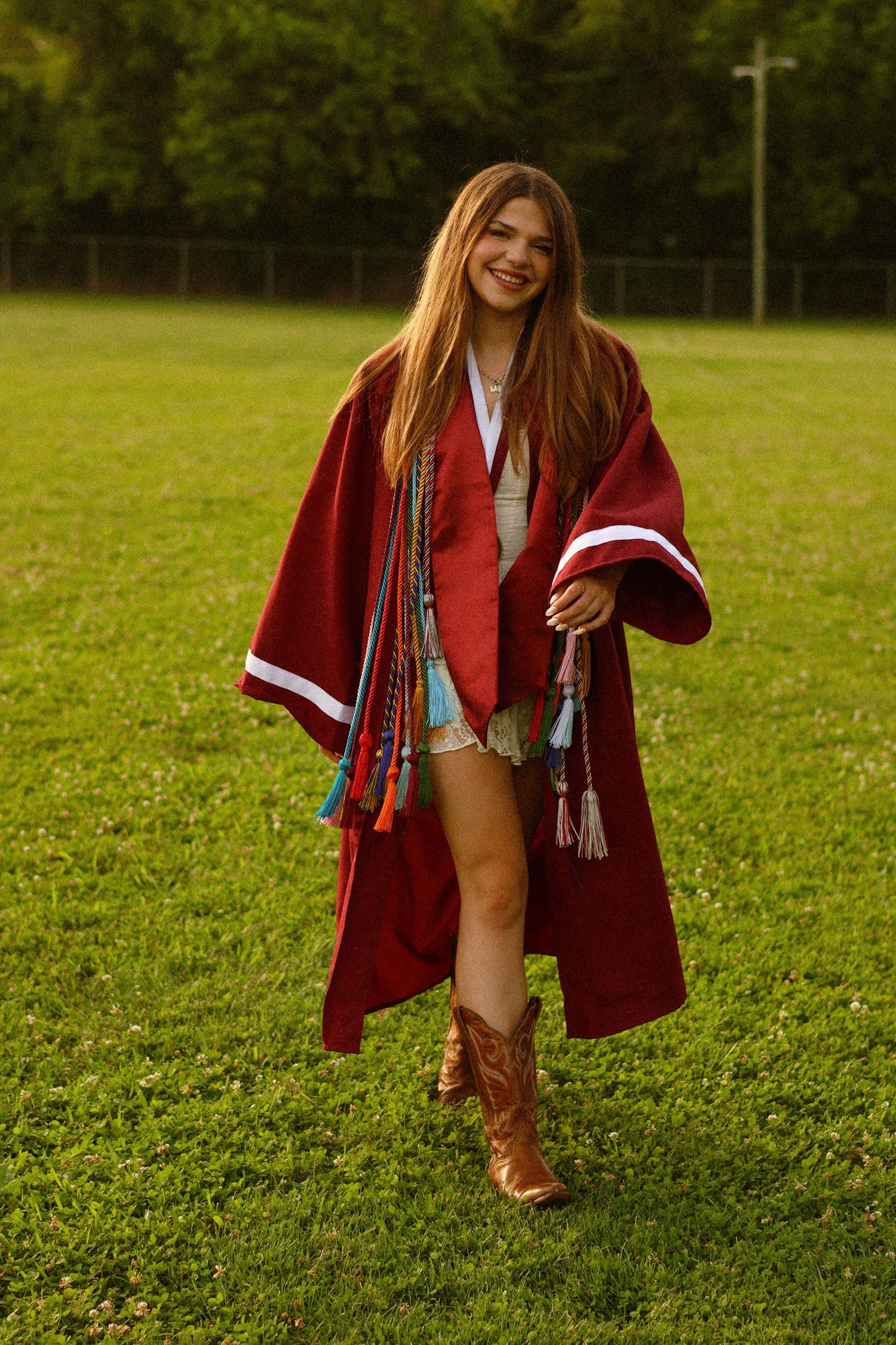 Smiling young woman in red graduation gown with colorful honor cords and cowboy boots standing on green grass.