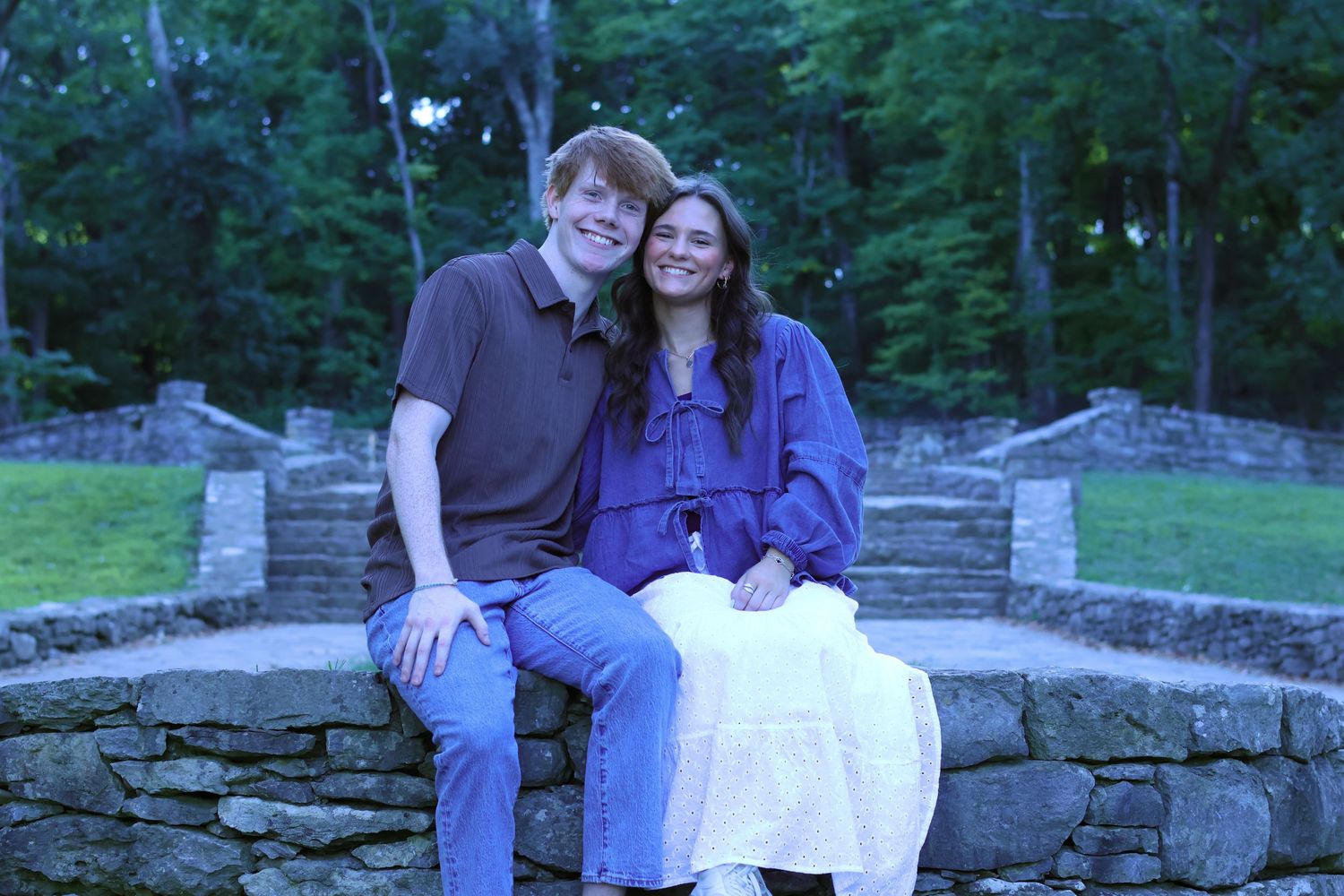 Couple relaxing on stone wall in wooded park setting.