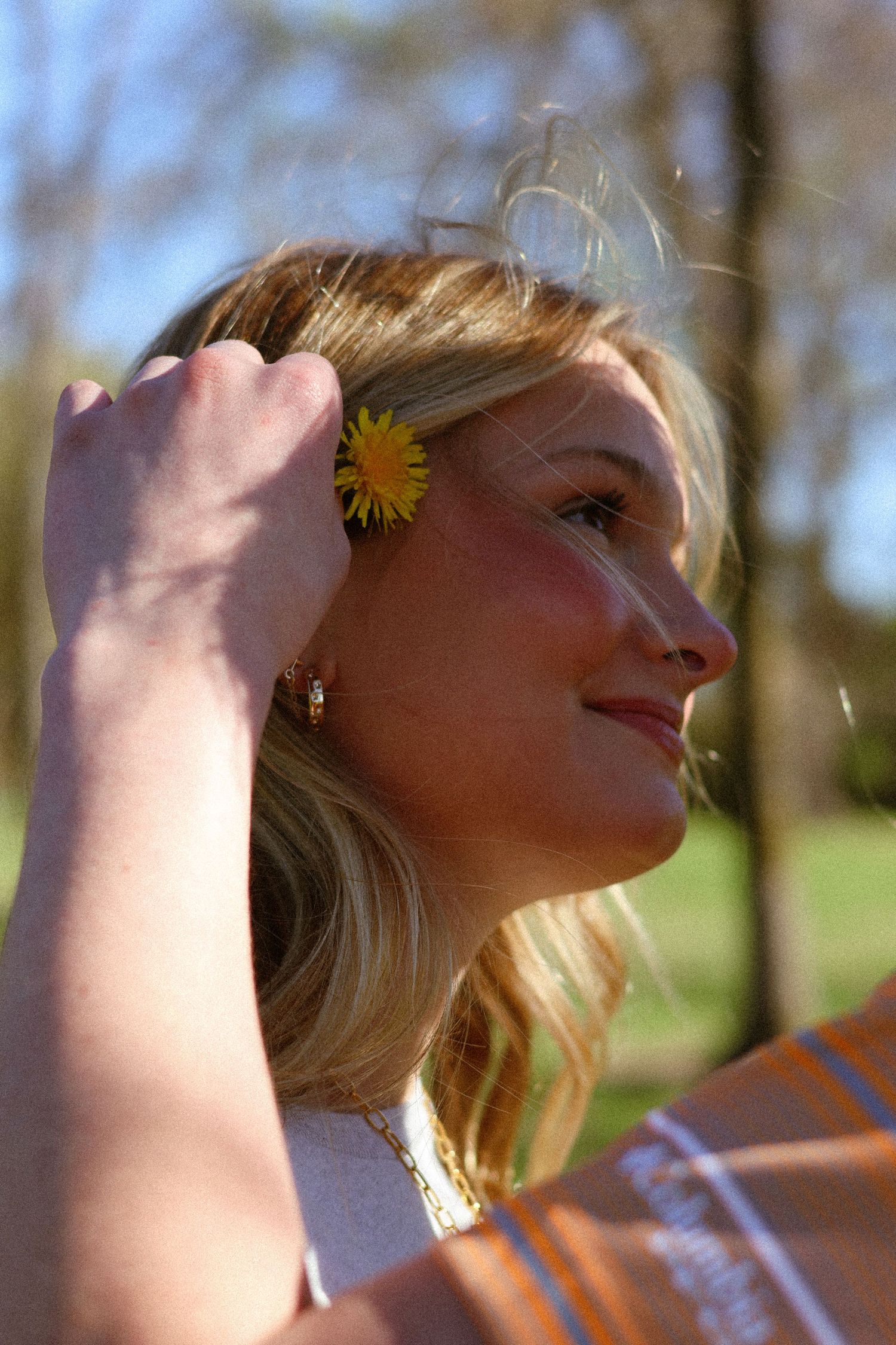 Young blonde woman smiling outdoors with a yellow dandelion flower tucked behind her ear on a sunny day.