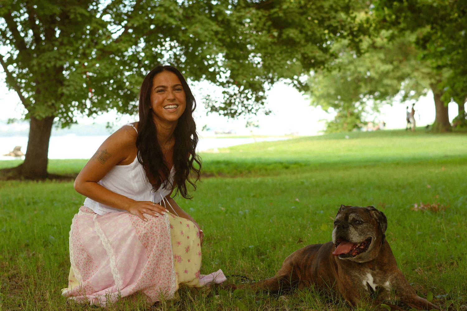 A smiling woman in a pink skirt sits on green grass beside a brown pit bull dog under shady trees in a park.