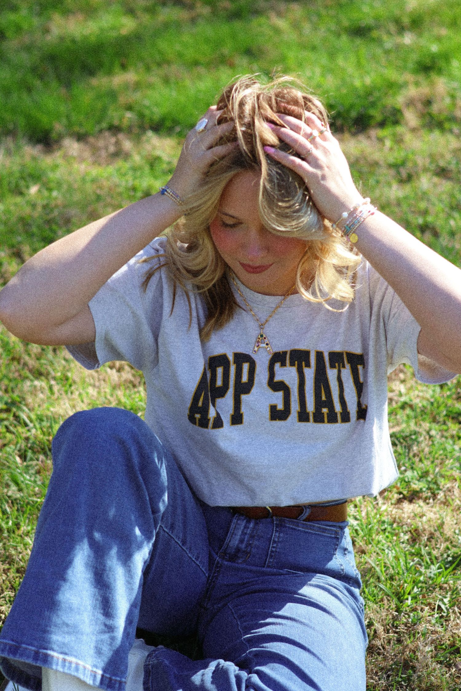 Blonde woman sitting on grass wearing an App State t-shirt and jeans, hands in hair.