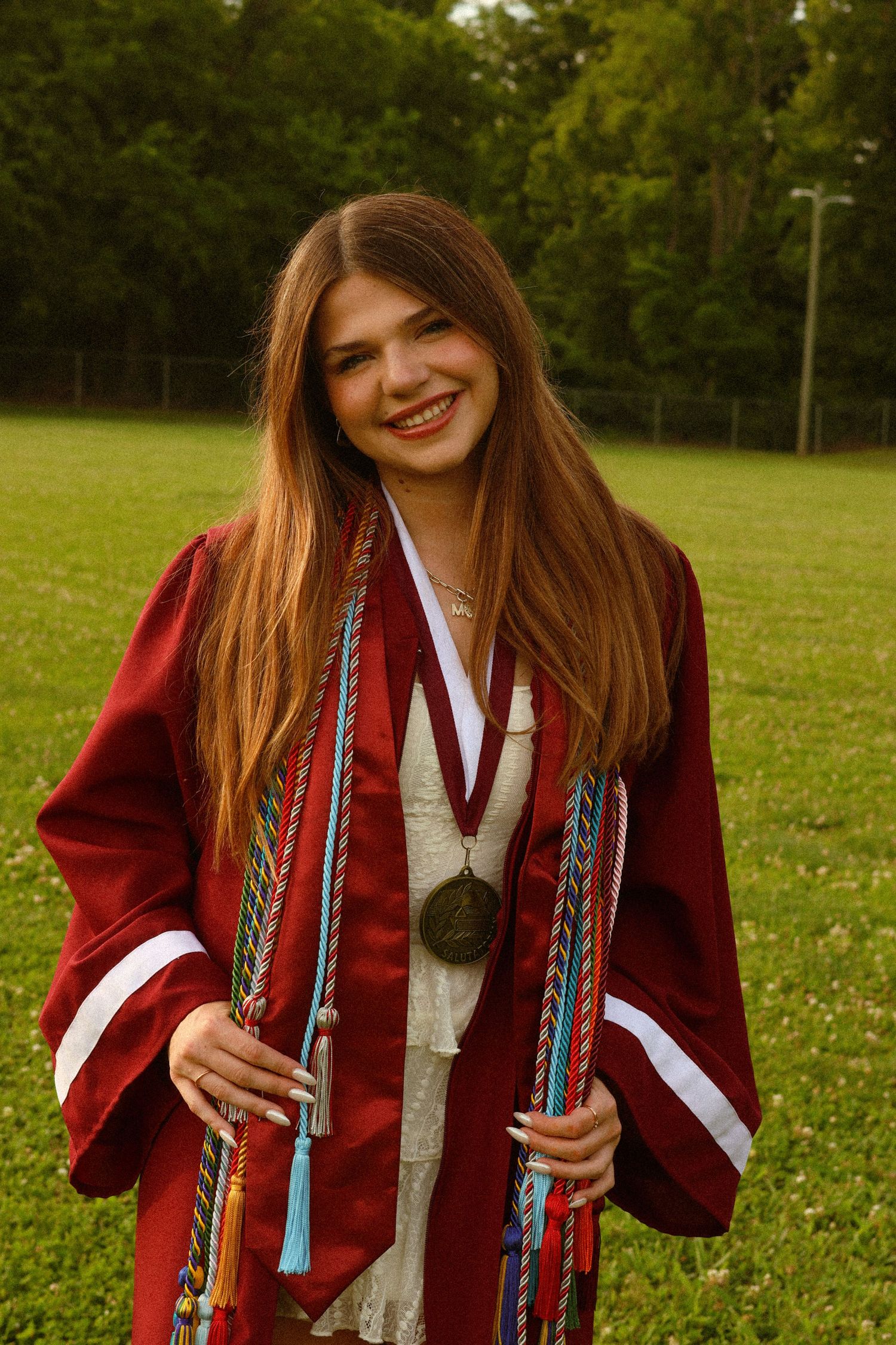 Smiling young woman in red graduation gown with colorful honor cords and medal posing outdoors on green grass.