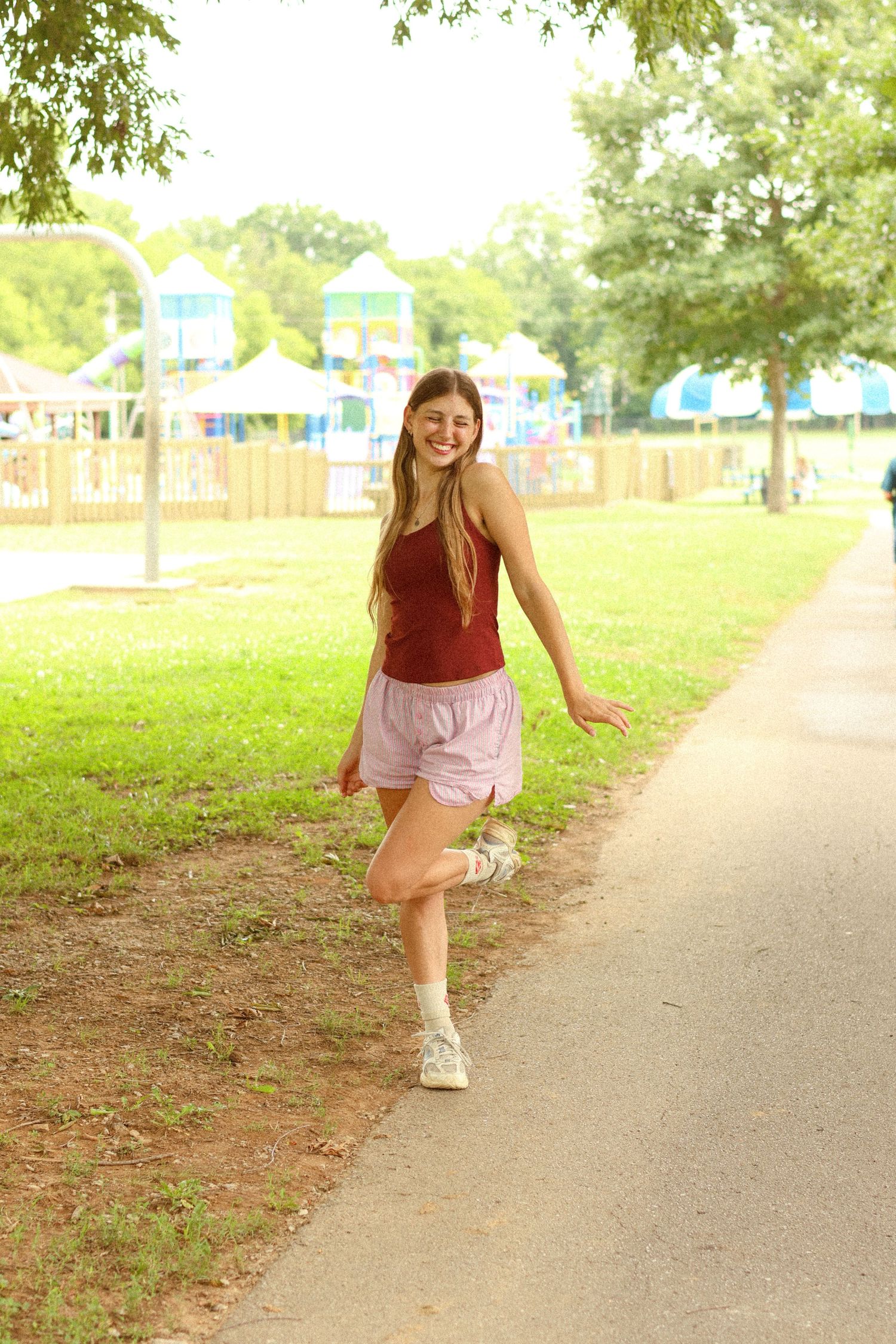 Young woman in red top and pink shorts smiling and walking on a park path near a colorful playground.