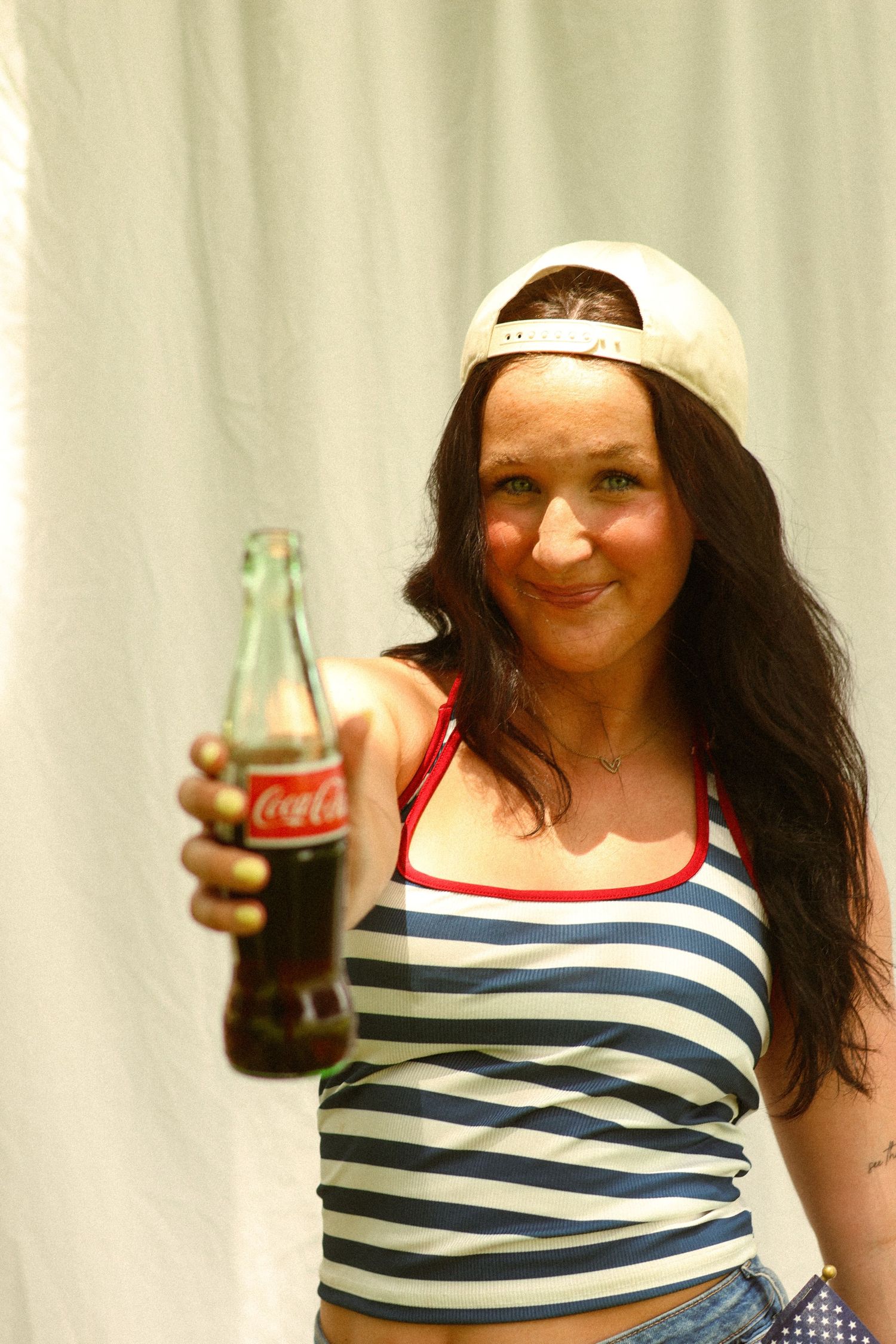 Woman in striped crop top extending Coca-Cola glass bottle toward camera, wearing white cap backwards.