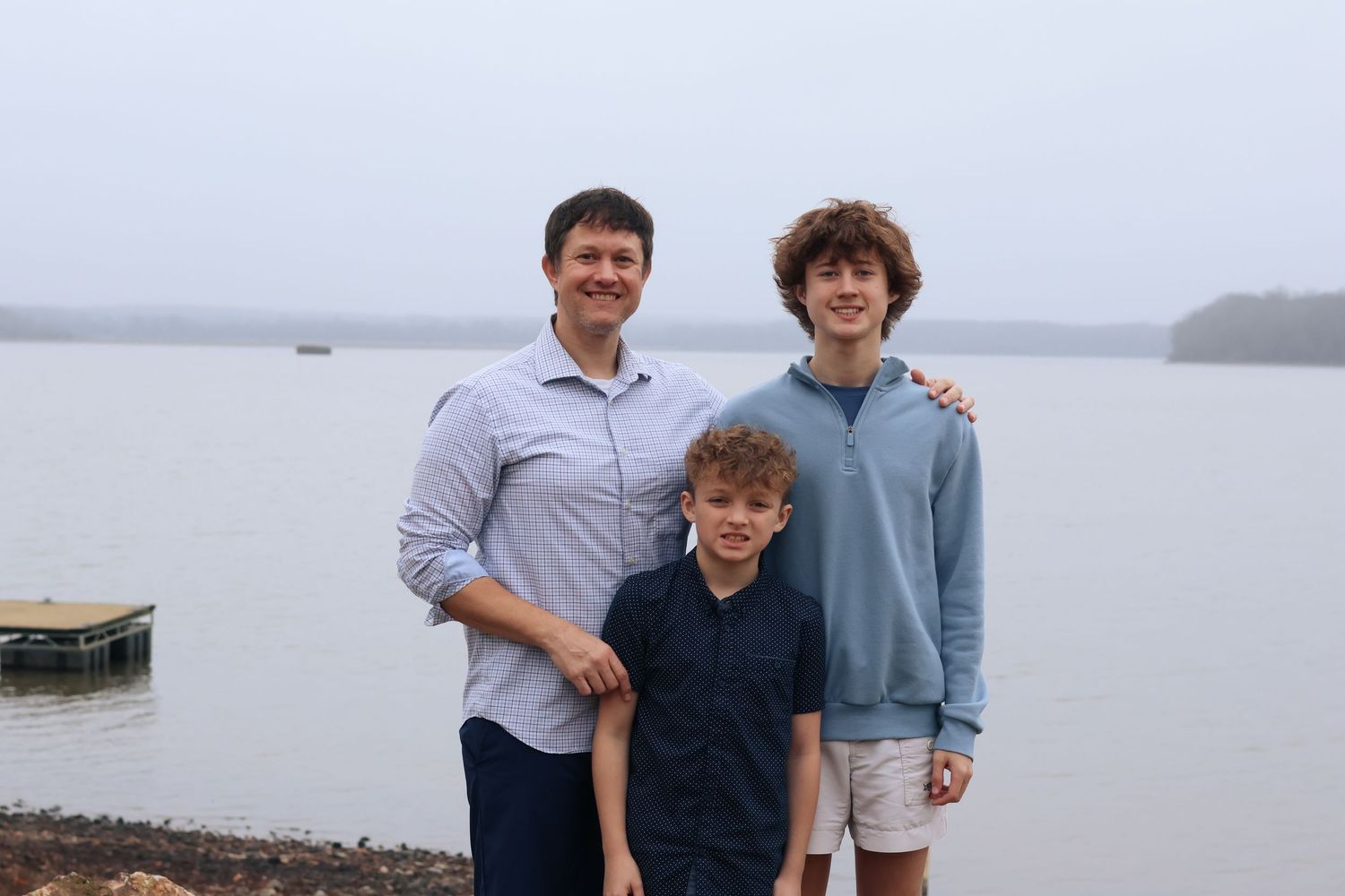 Three people pose together on a dock by a foggy lake on a cloudy day.
