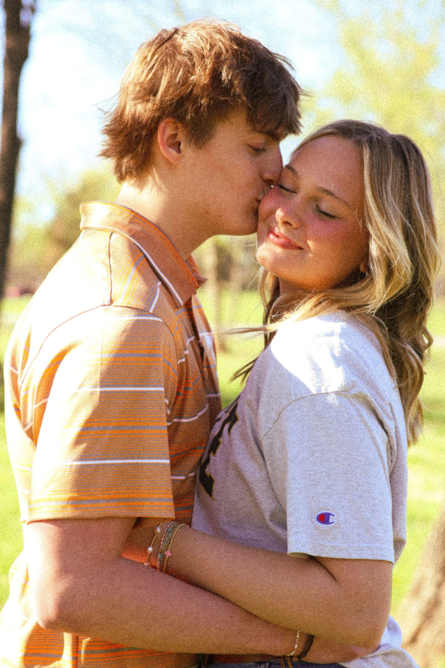 A young couple shares a tender kiss outdoors, with the boy in an orange striped shirt and girl in a Champion tee.