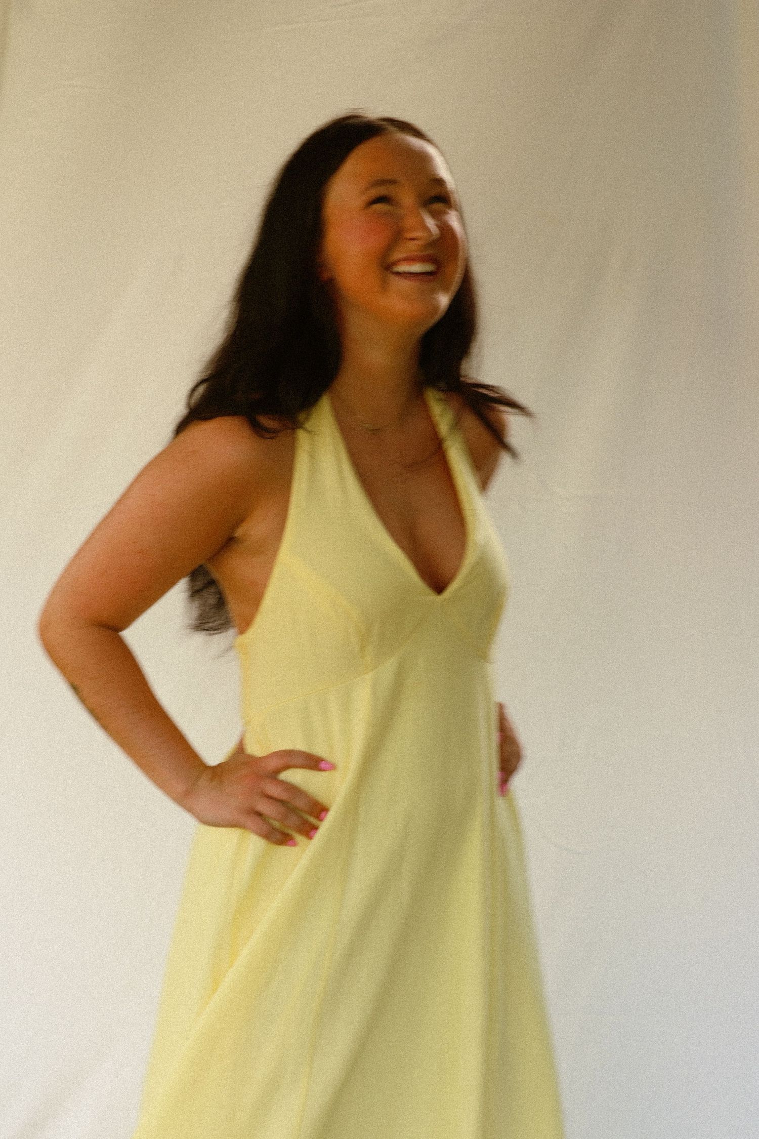 Woman in a pale yellow halter dress posing with hand on hip, laughing against a white fabric background.