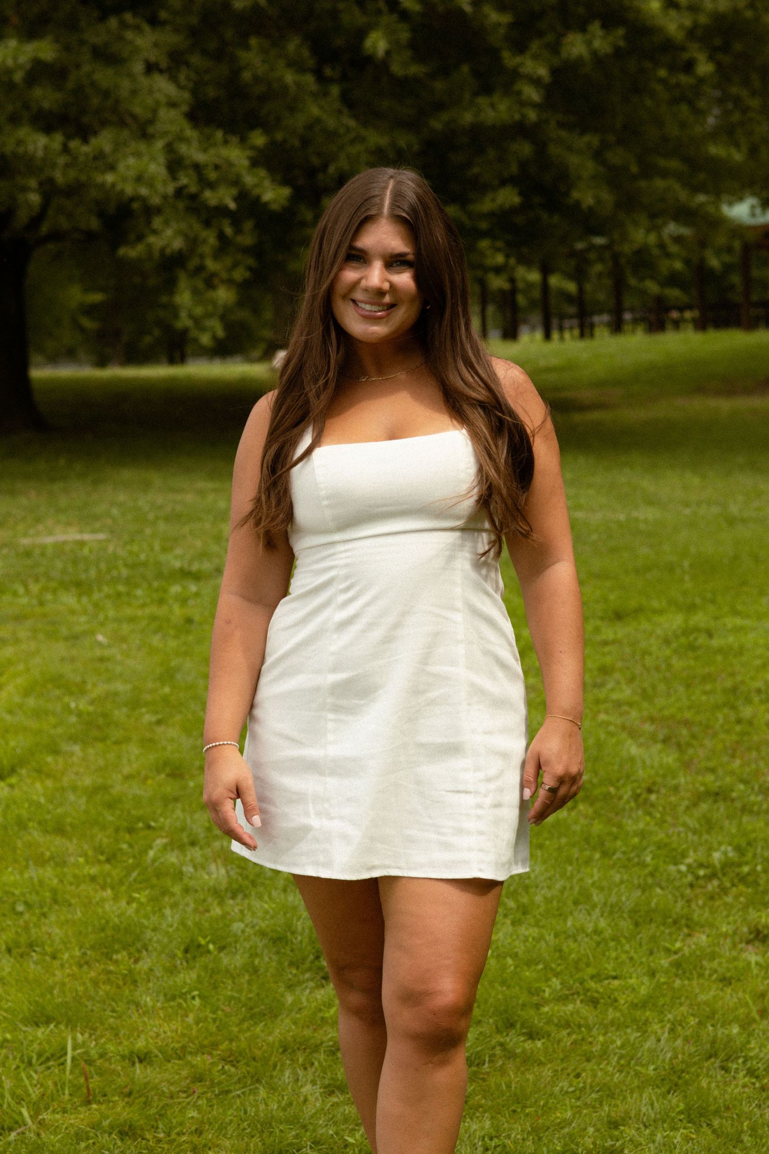 Smiling young woman in a white strapless mini dress standing on green grass in a park on a sunny day.