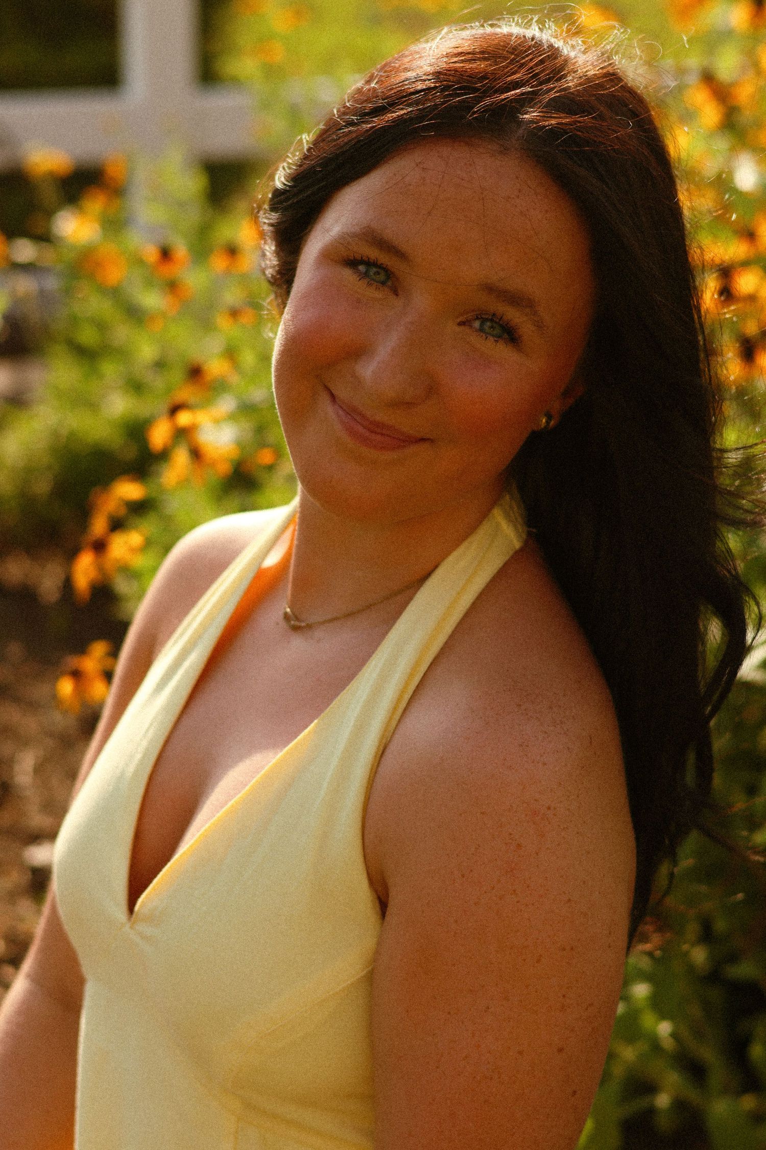 A young woman with dark hair and freckles smiles outdoors in a yellow halter dress, surrounded by orange flowers.
