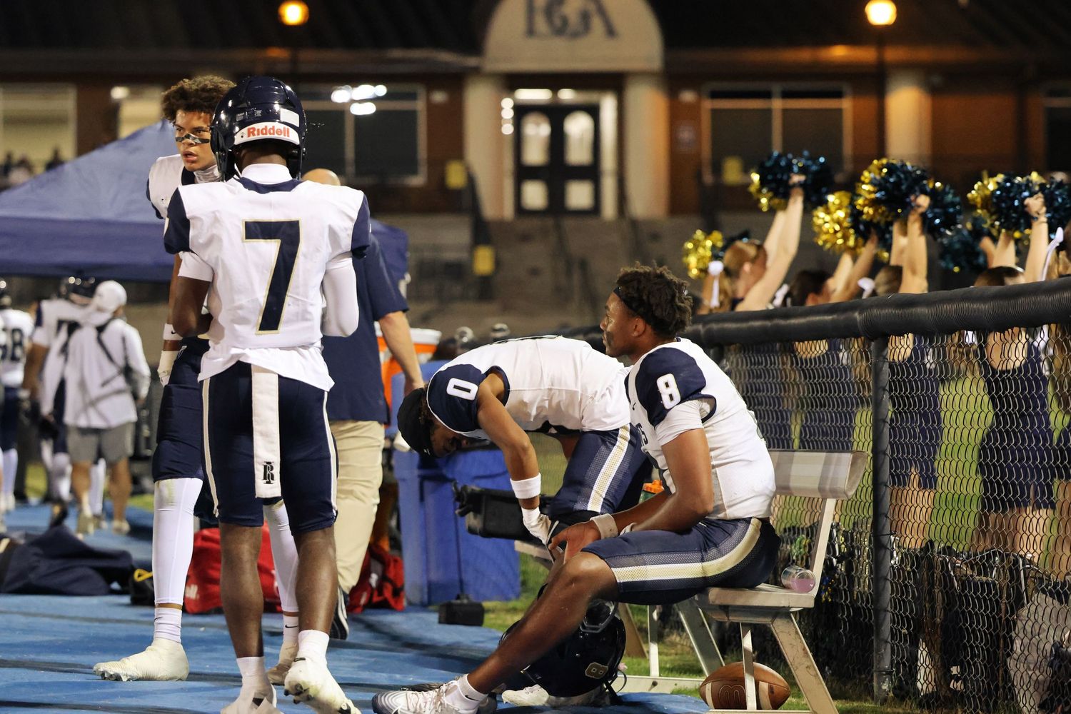 Football players and coaches gather on the sidelines during a night game.