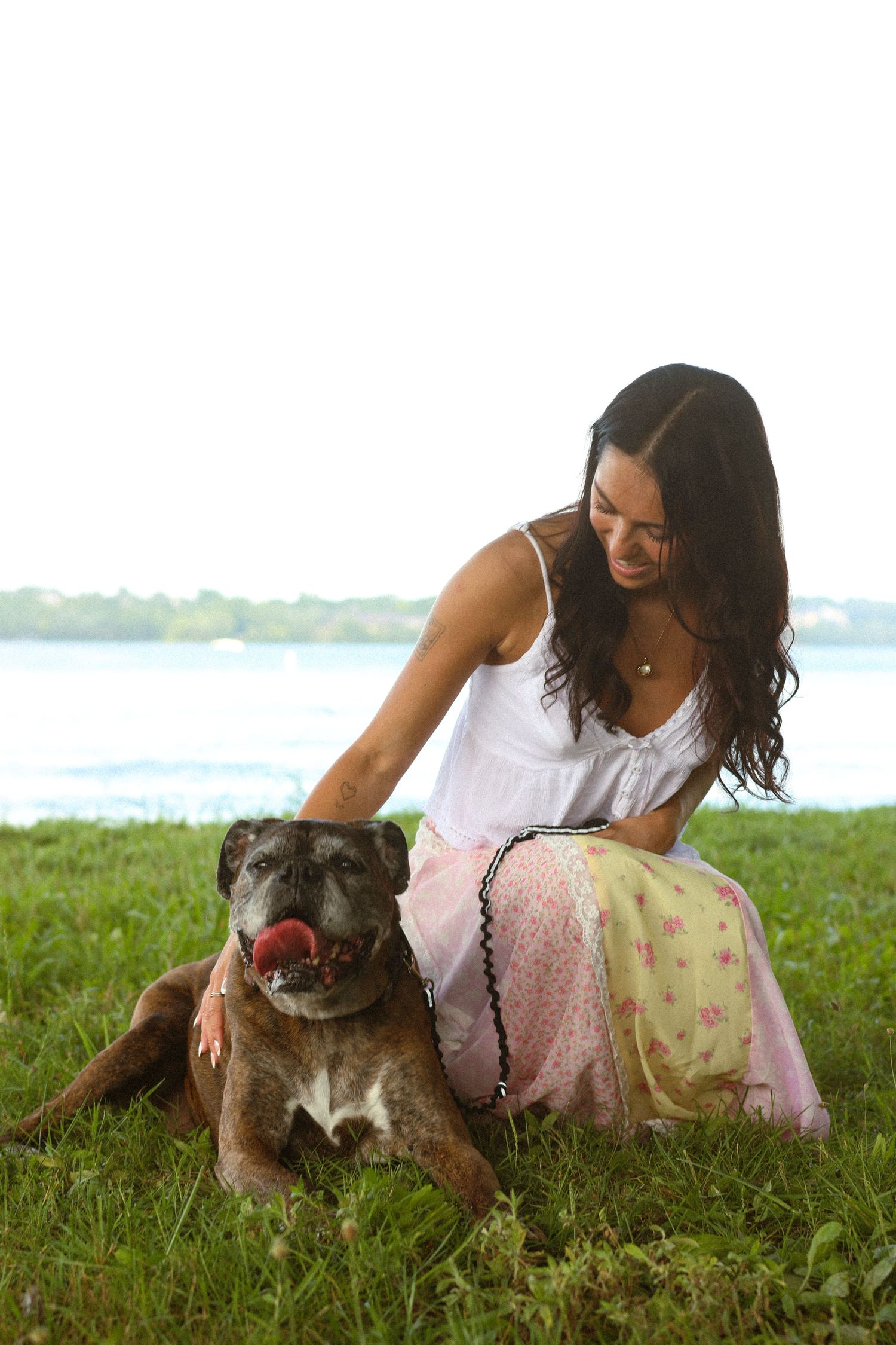 Woman in white top and pink skirt petting a brown dog on green grass near a lake.