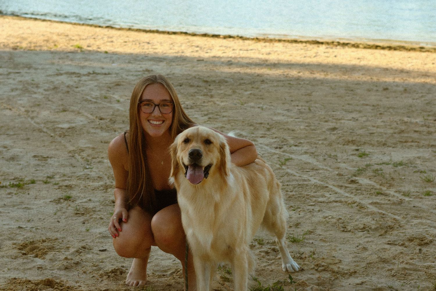 A smiling young woman with glasses crouches beside a golden retriever on a sandy beach near calm water.