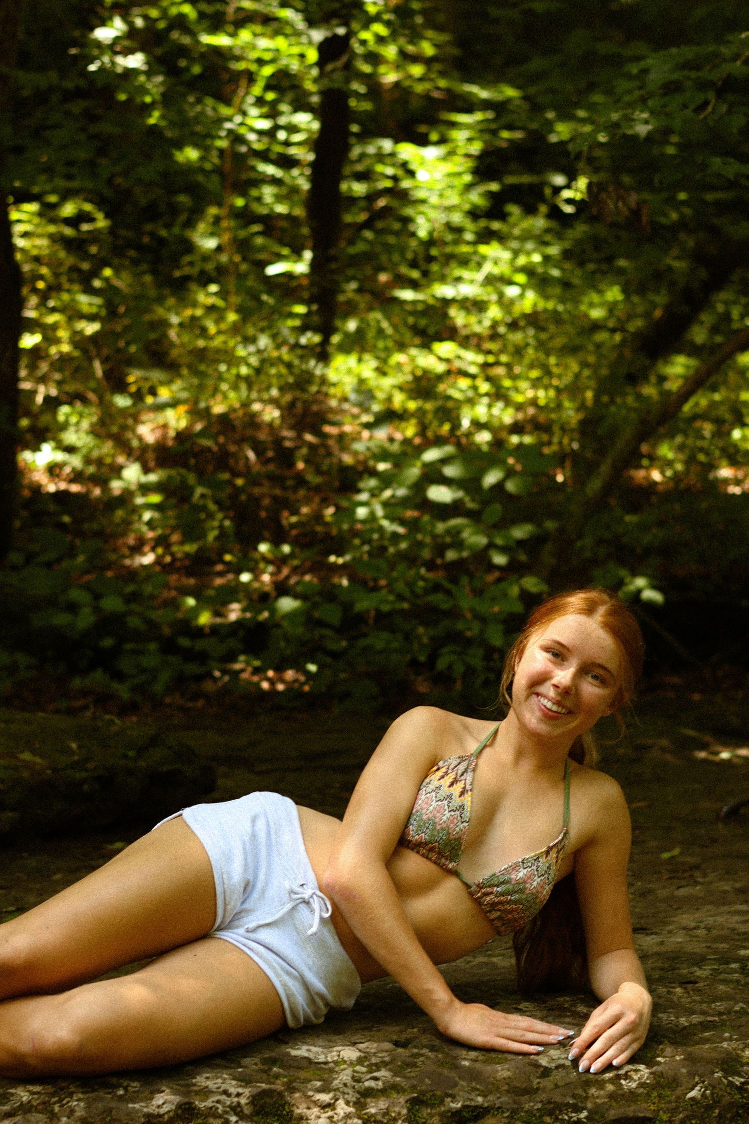 A young redhead woman in a bikini top and white shorts relaxing on a rock in a lush green forest.