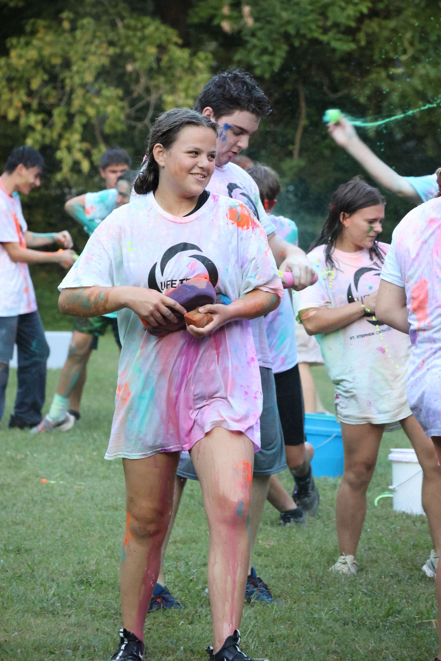 People in white t-shirts covered in colorful powder participate in an outdoor color run event on a grassy field.
