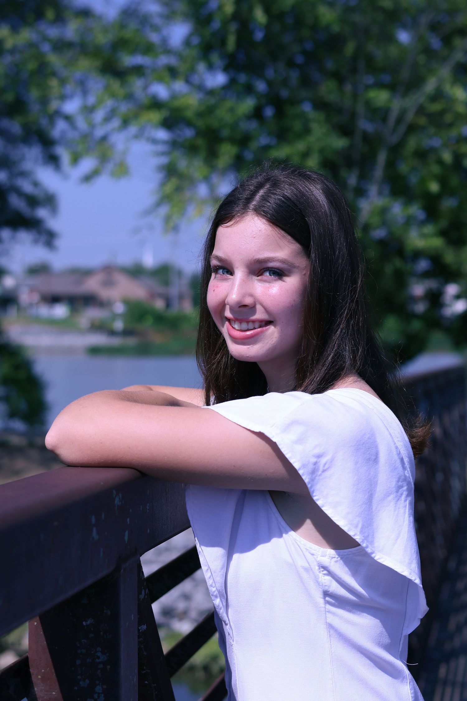 Young woman in white shirt poses playfully against outdoor railing with trees and water in background.