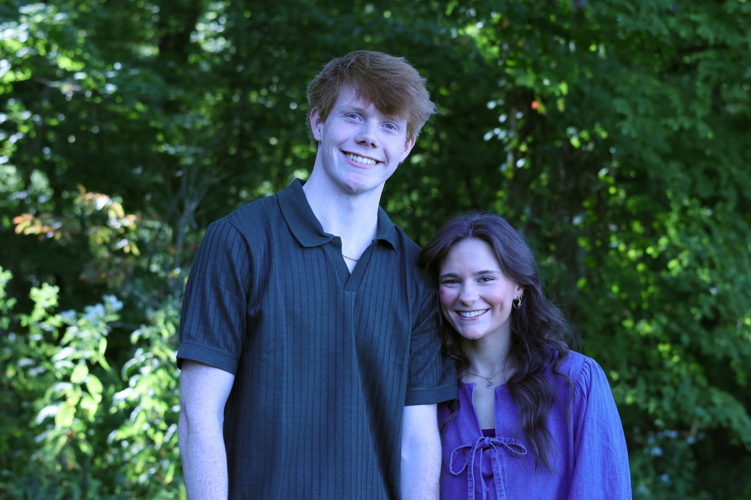 Couple standing together outdoors in casual attire against green foliage background.
