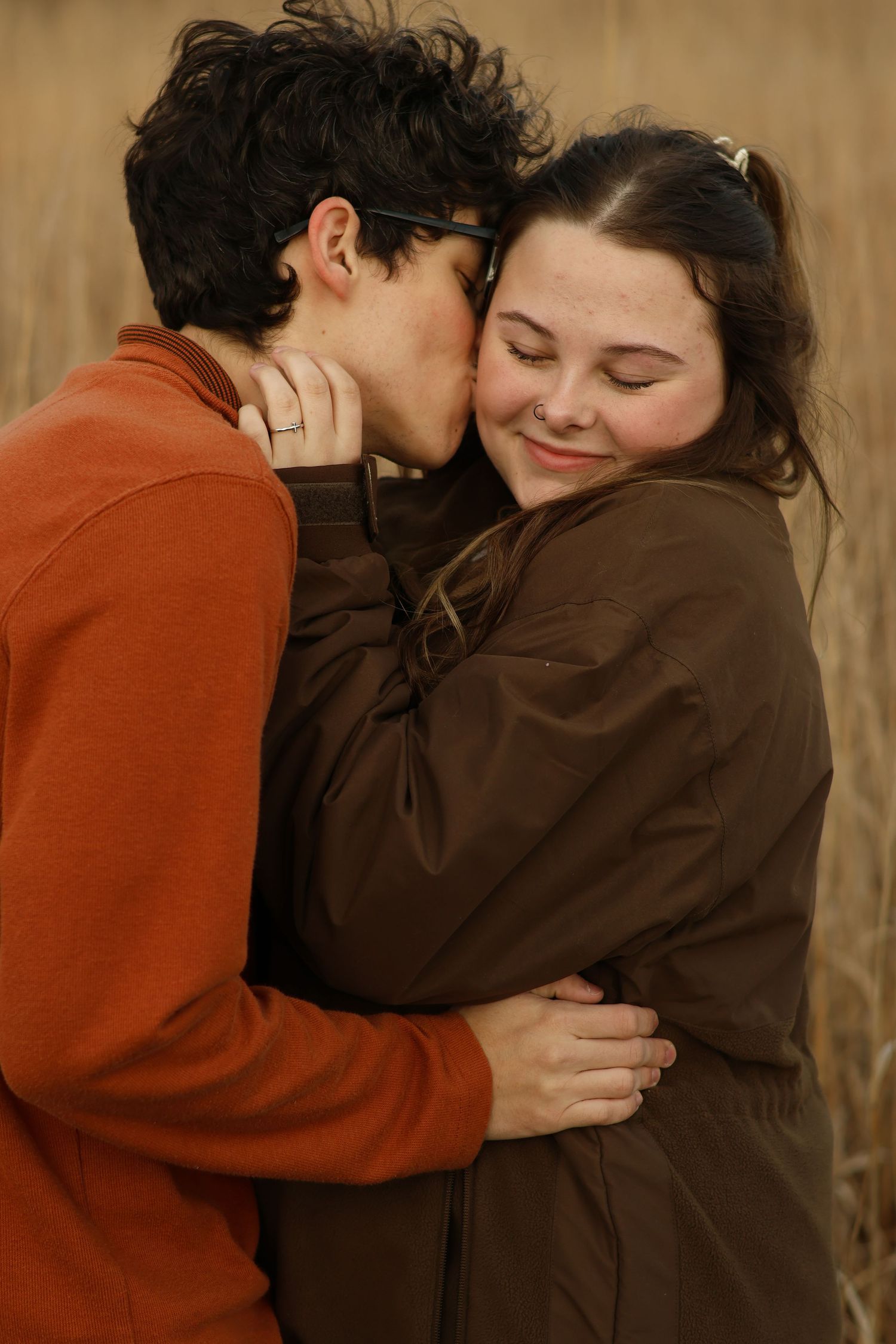 Two people in brown and red clothing embrace in a tender moment against a neutral background.
