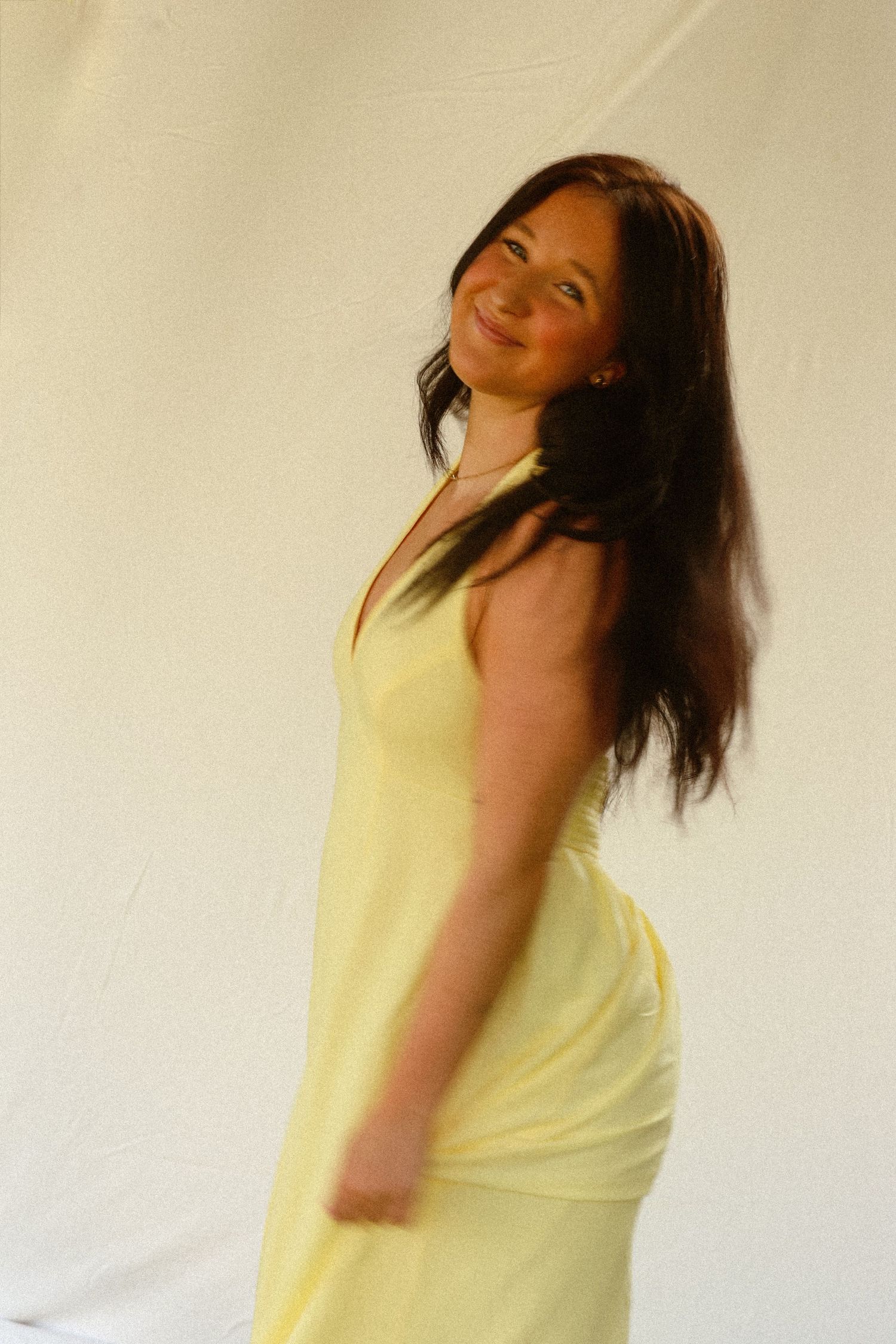 Smiling woman with long dark hair wearing a light yellow halter dress, posing against a white backdrop.