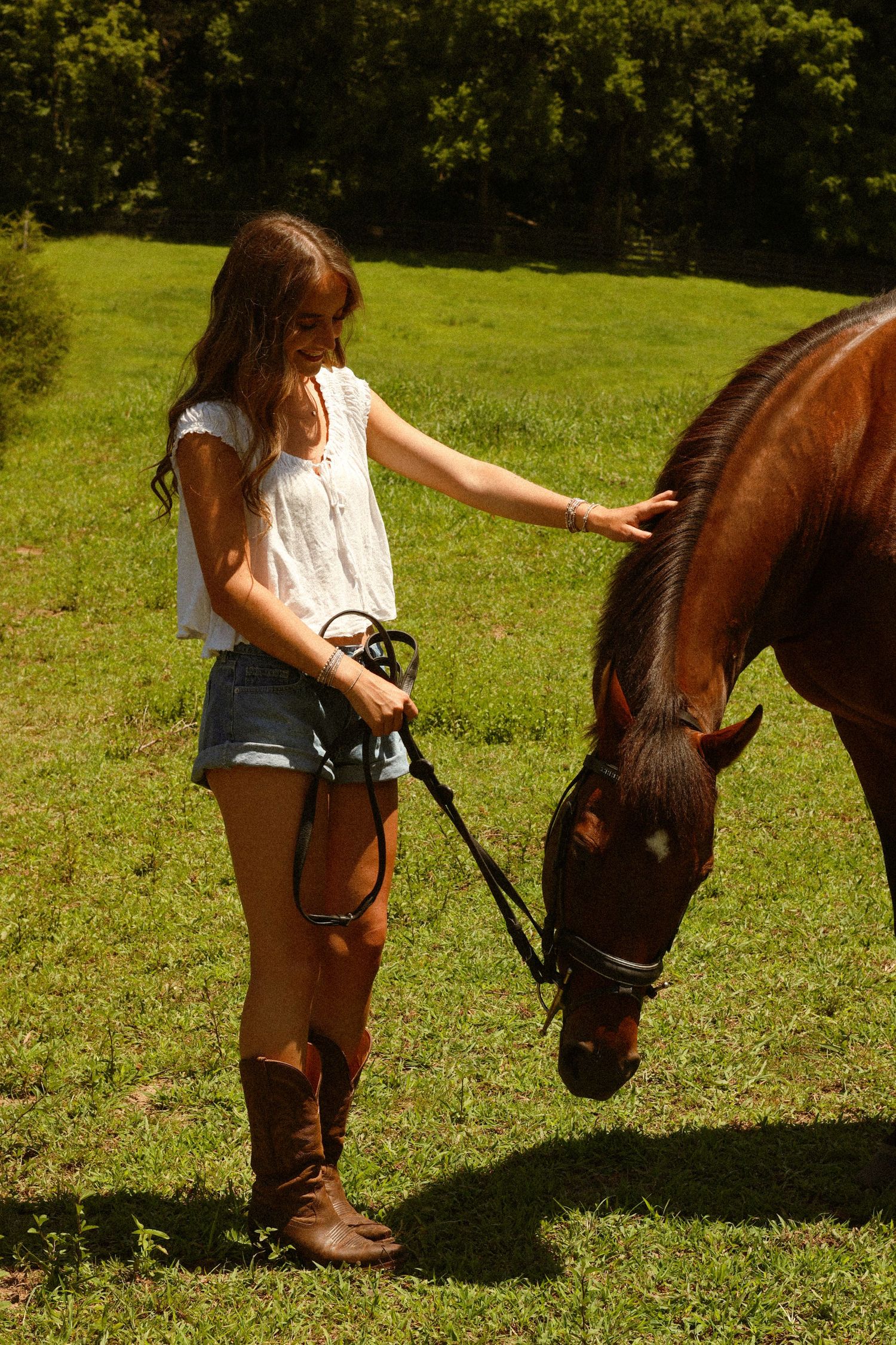 A young girl in denim shorts and boots gently pets a brown horse on a sunny green pasture.