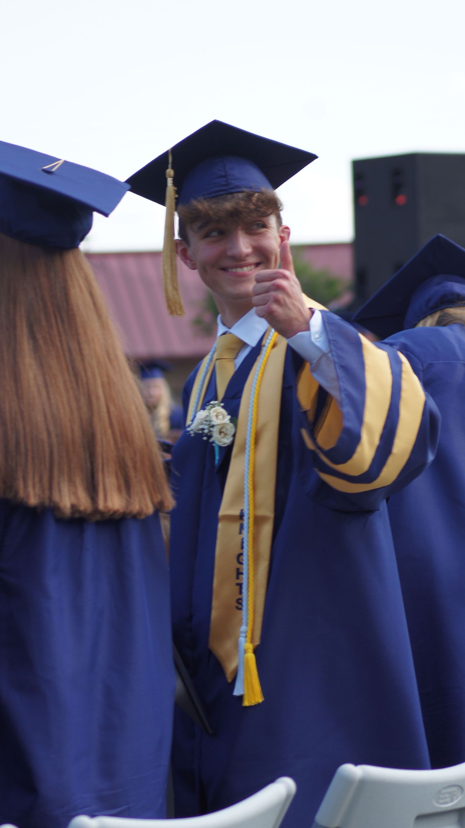 Graduates in blue caps and gowns with gold honor cords stand together during commencement ceremony.