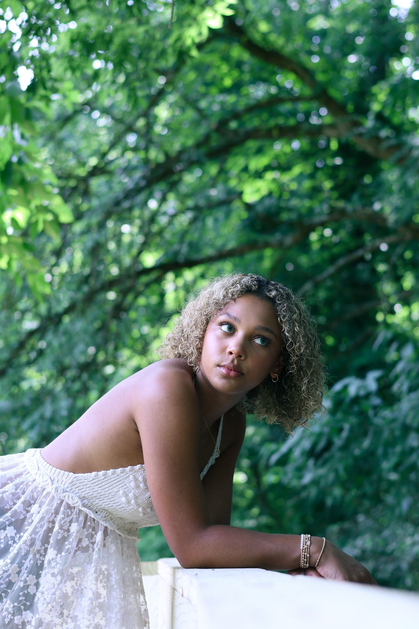 Person in white lace top poses elegantly against green forest backdrop.