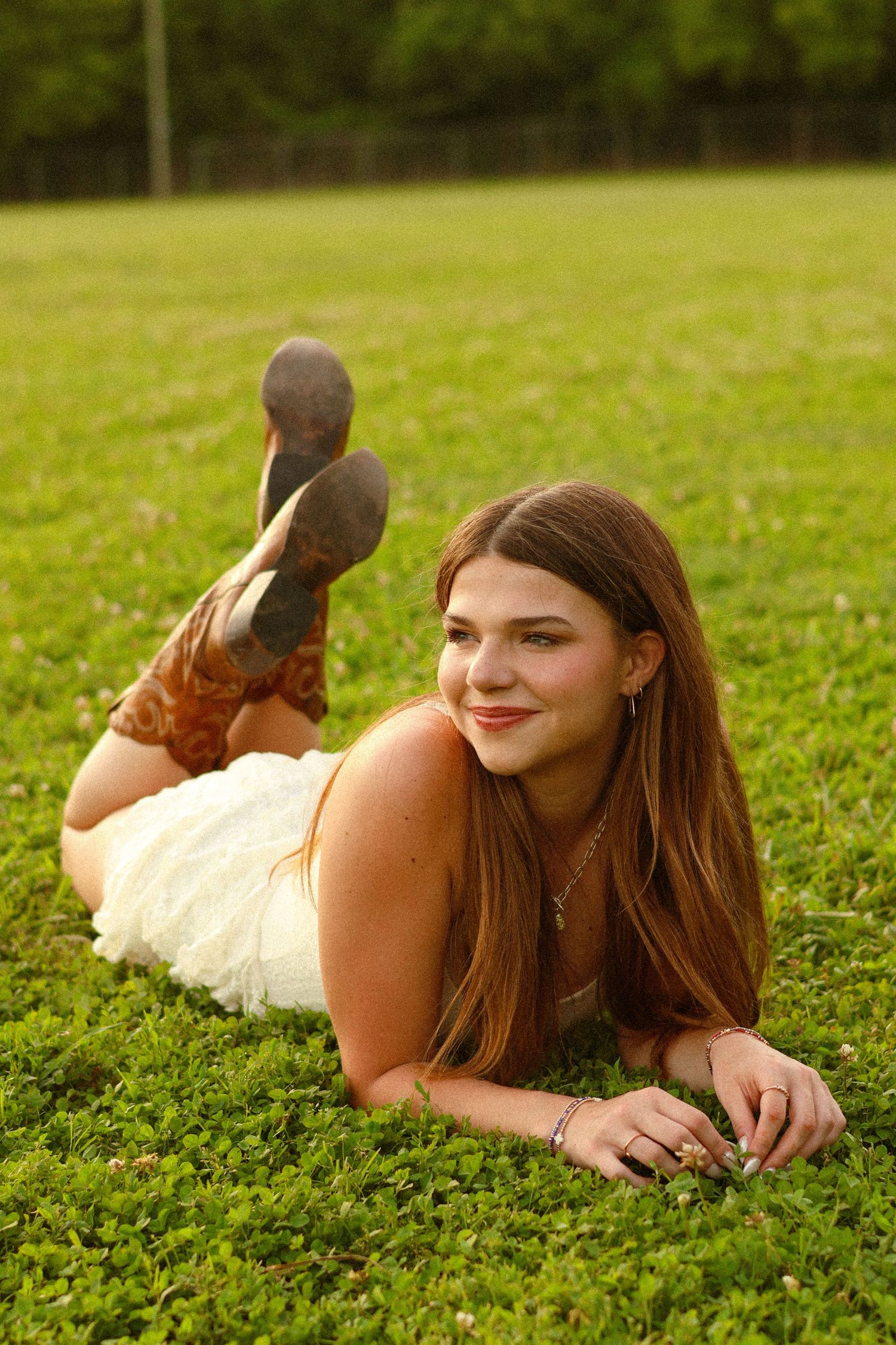 Young woman in white dress and cowboy boots lying on green grass, smiling outdoors in natural light.
