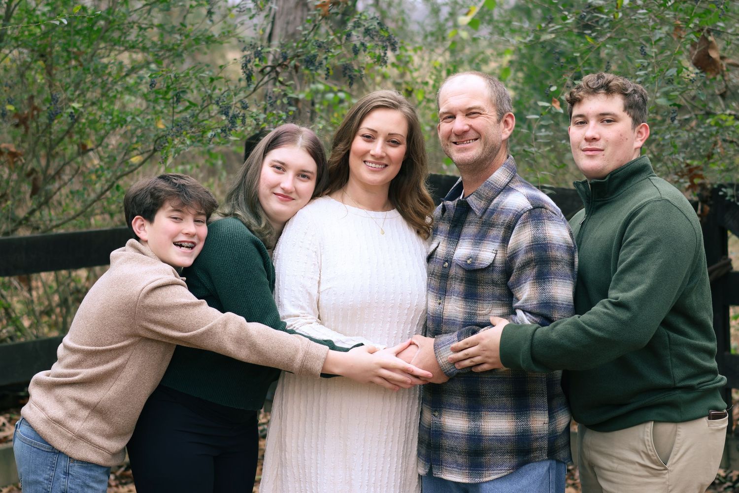 A group of people in casual winter attire pose together outdoors among trees and greenery.