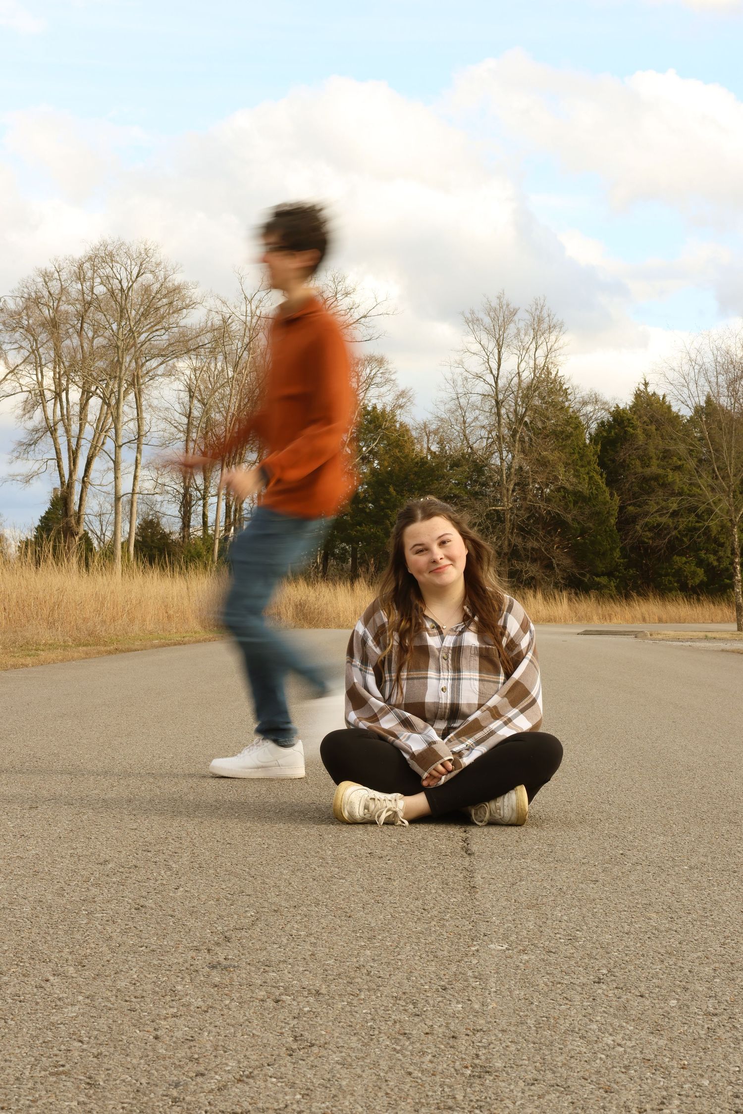 Two people on an empty road with bare trees and grass in the background, one sitting while another walks past in motion blur.