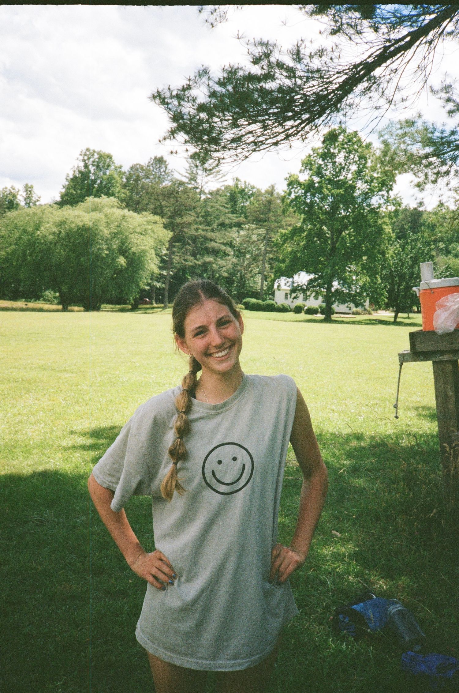 Someone wearing a grey t-shirt with smiley face design poses outdoors in a grassy park setting.