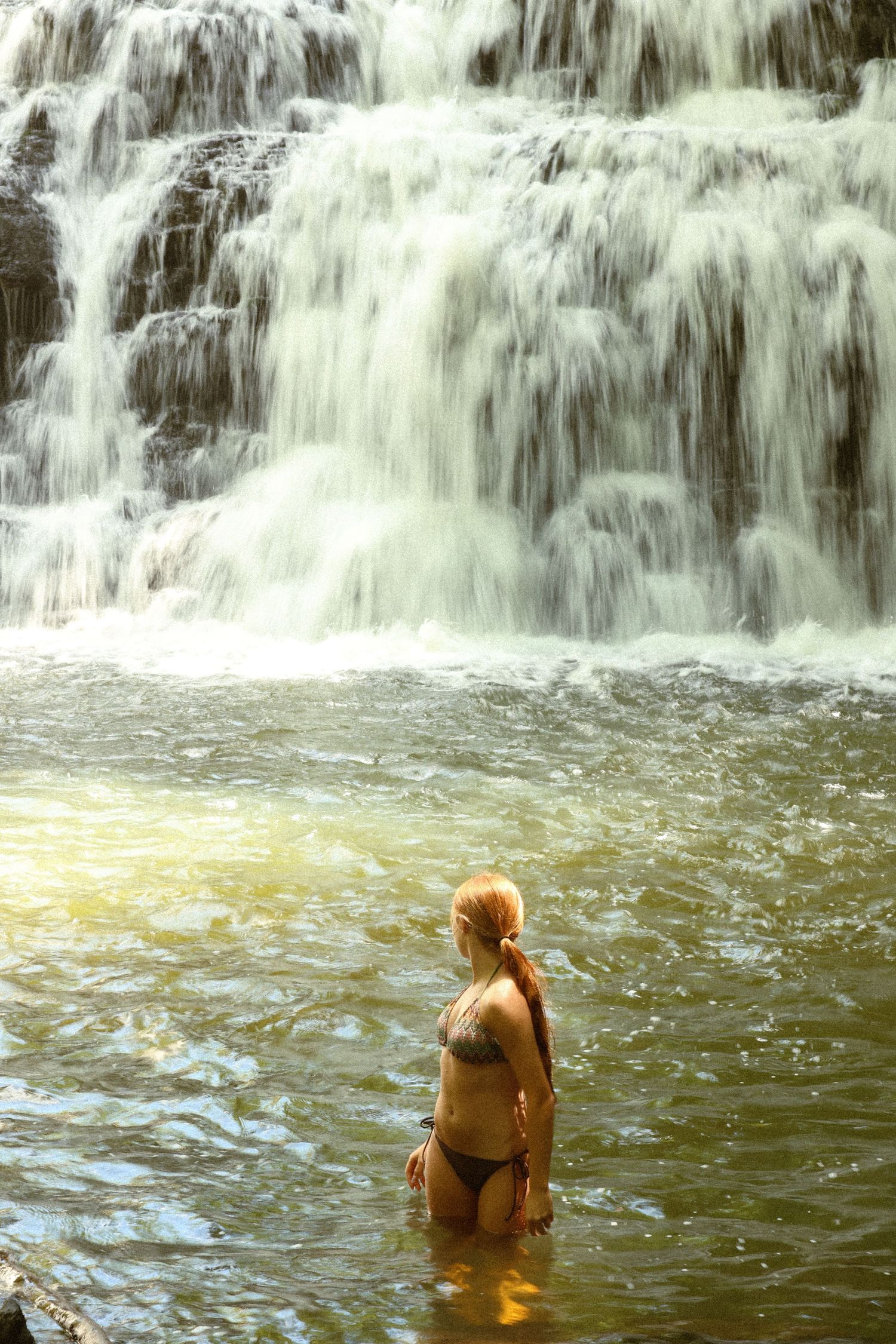 Woman in bikini standing waist-deep in a natural pool before a stunning multi-tiered cascading waterfall.