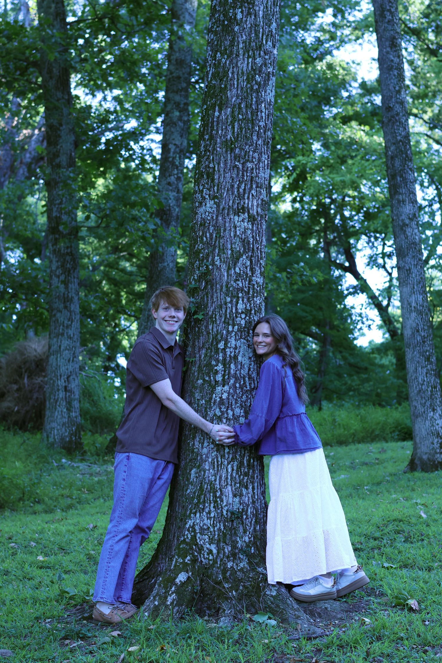 Couple poses playfully on opposite sides of large tree trunk in wooded setting.