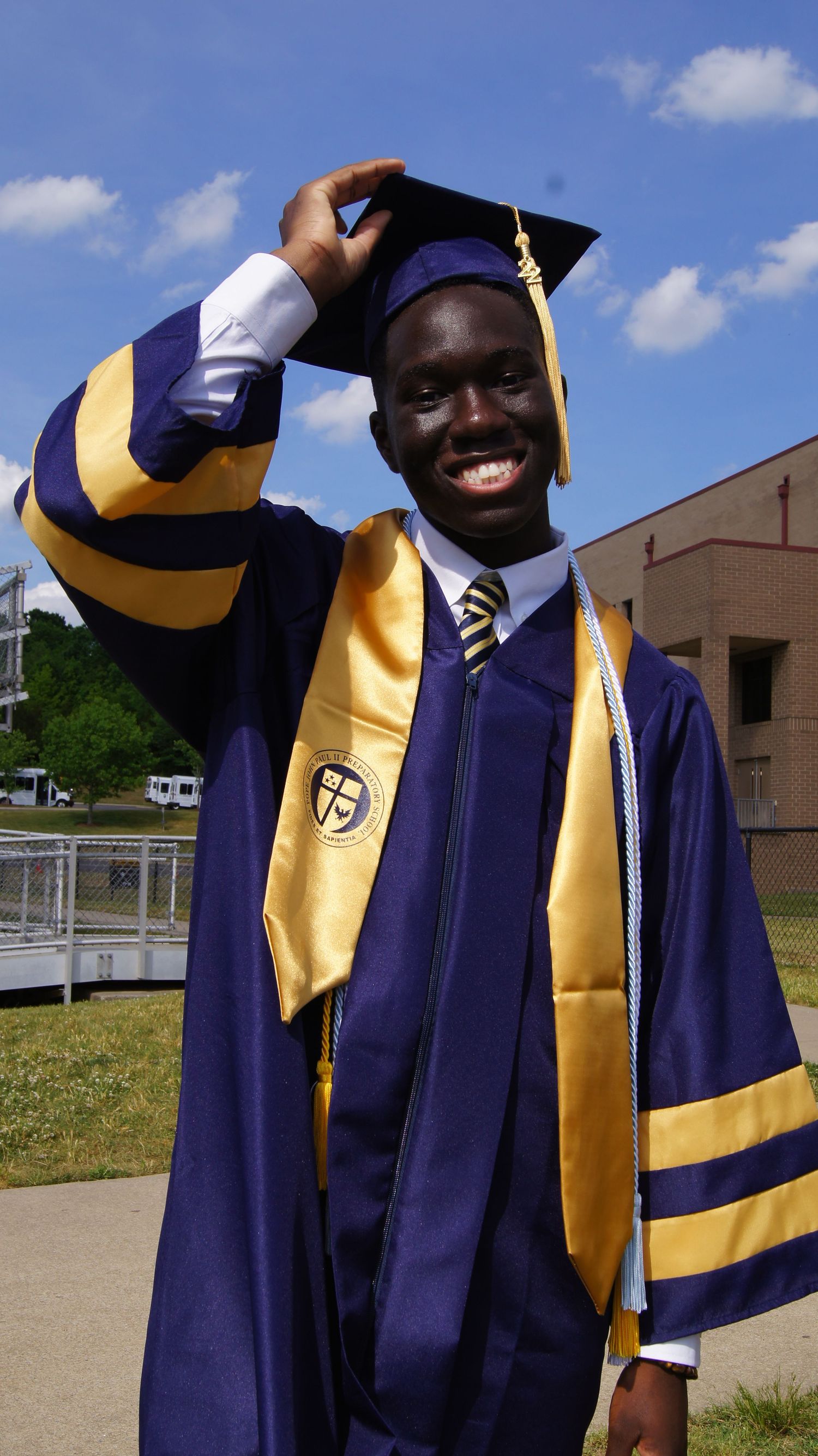 Graduate in navy and gold academic regalia celebrates achievement with joyful poses outdoors on sunny day.