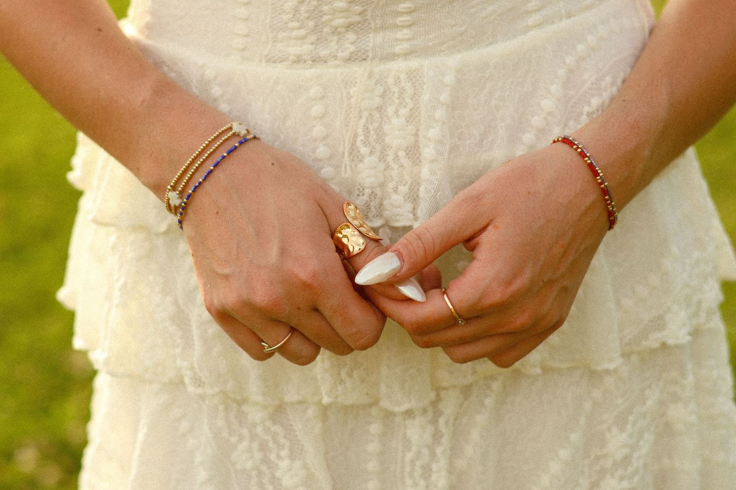 Close-up of woman's hands adorned with colorful bracelets and rings, clasped together over a white lace dress.