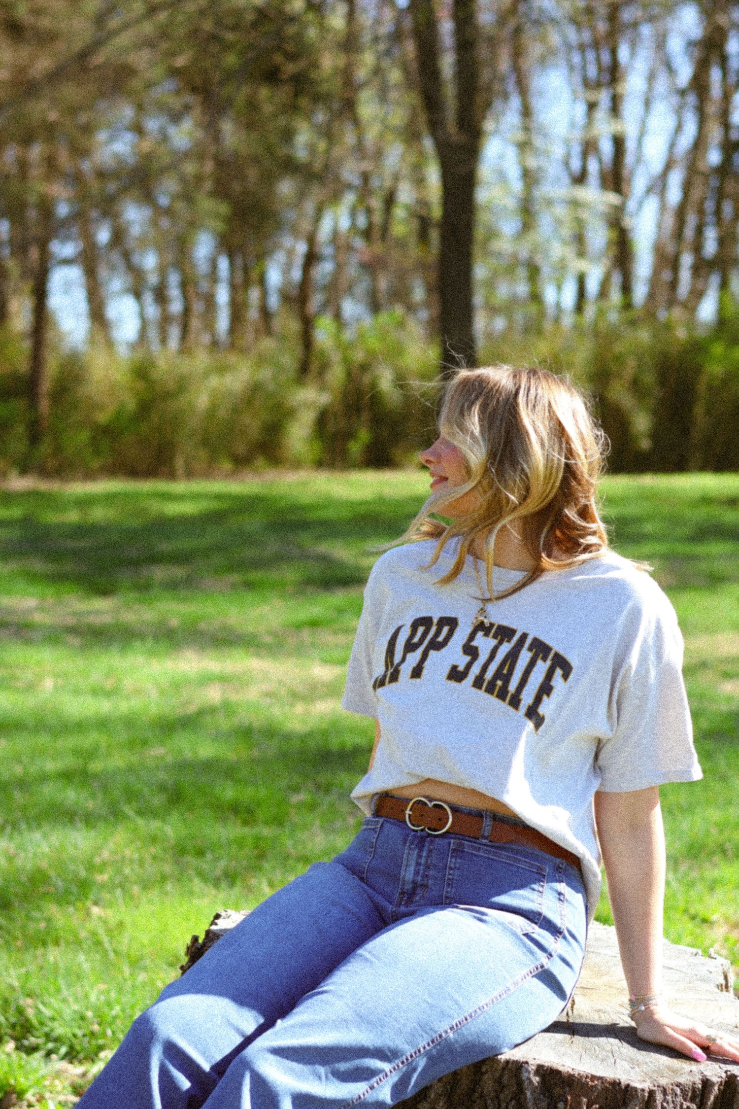 Woman in App State t-shirt and jeans sitting outdoors on sunny day with green grass and trees.