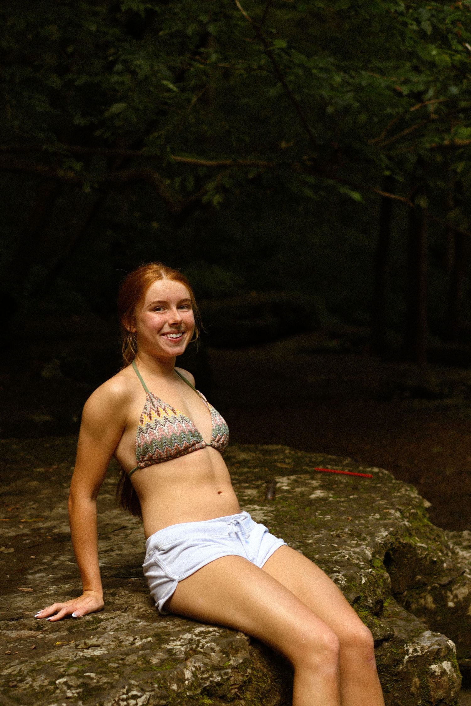 Young woman with red hair in bikini top and white shorts sitting on rocks in a forest setting, smiling.