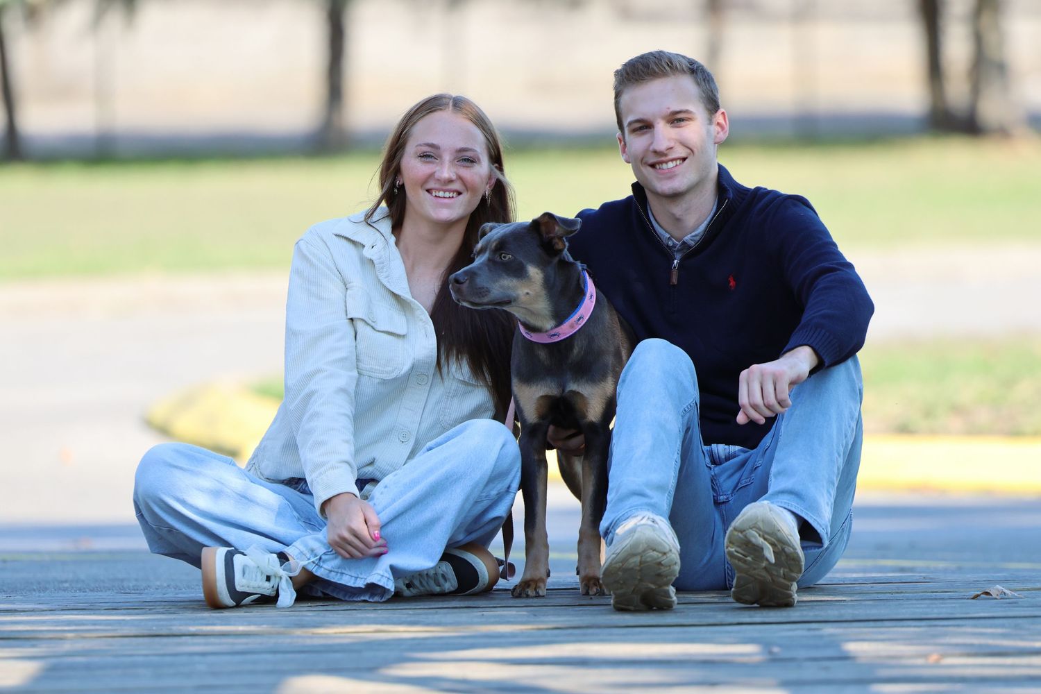 A couple sits with their black dog on a wooden deck outdoors.