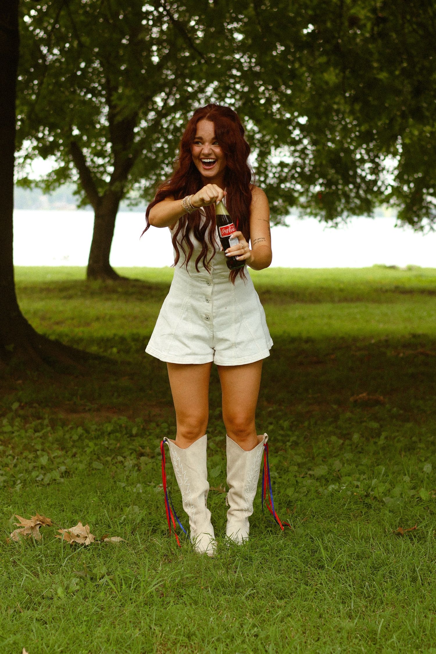 Young woman with red hair in white romper and cowboy boots opening a Coca-Cola bottle outdoors.