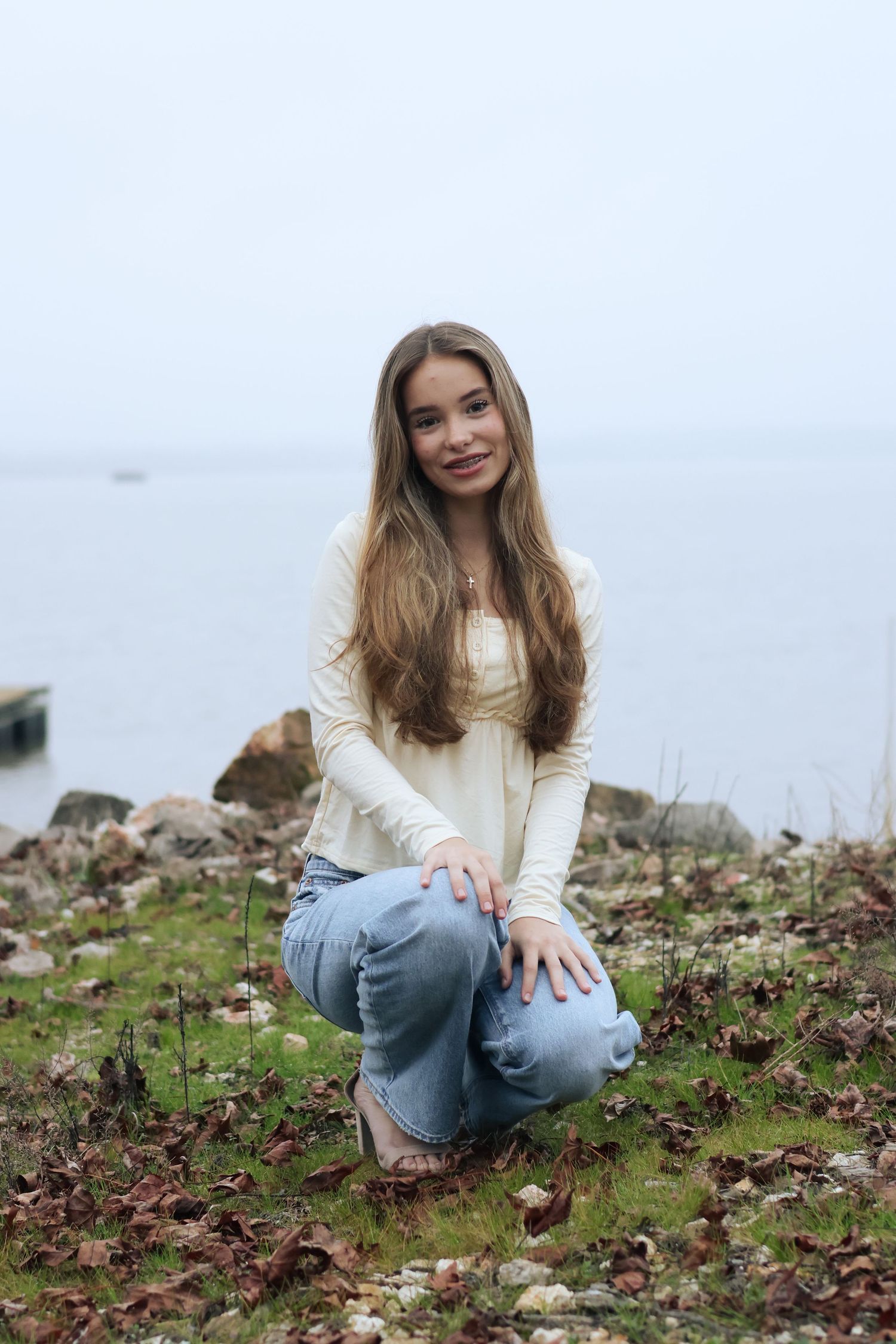 Person in light sweater and denim sits among wildflowers on coastal terrain during outdoor portrait session.