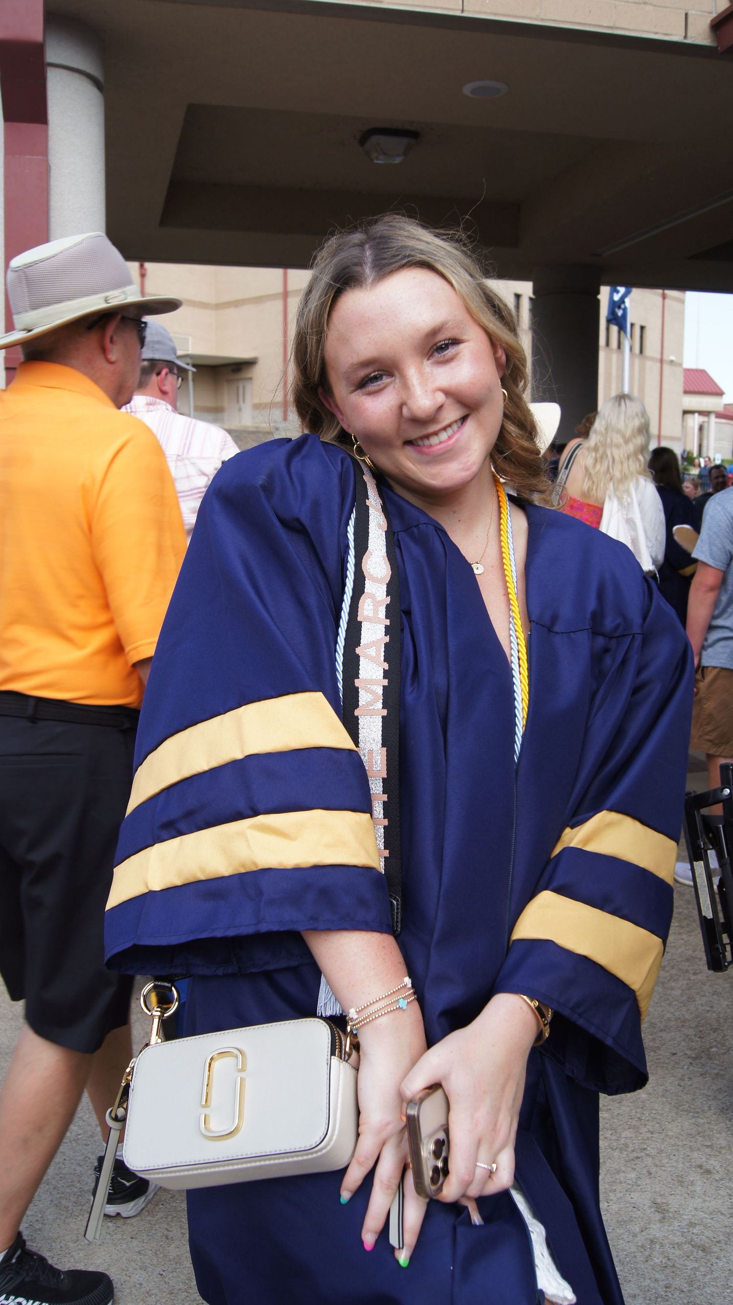 A person in graduation attire shows off their navy and gold gown while carrying a white purse.