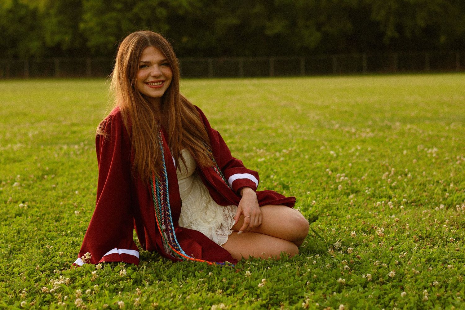 Young woman with long hair sits cross-legged on green grass, smiling, wearing a red jacket and white dress.