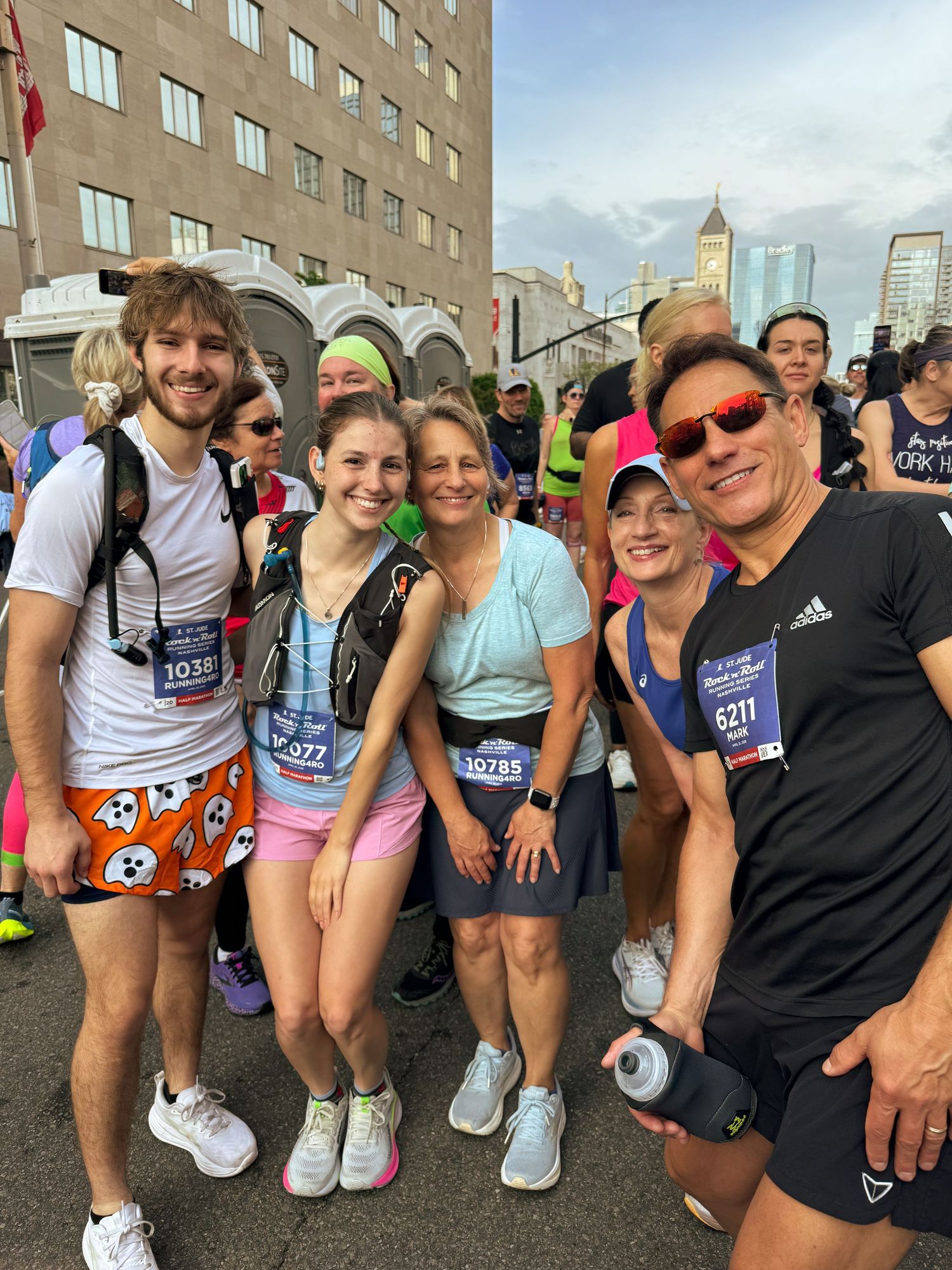 Group of runners posing together at a city marathon race, wearing race bibs and athletic gear, with city skyline in background.
